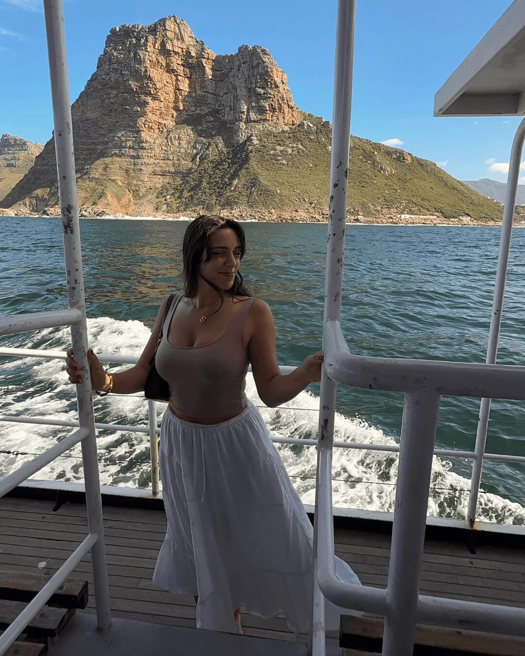 Woman standing on boat with mountain and water in background, holding onto boat railing, smiling, sunny day.