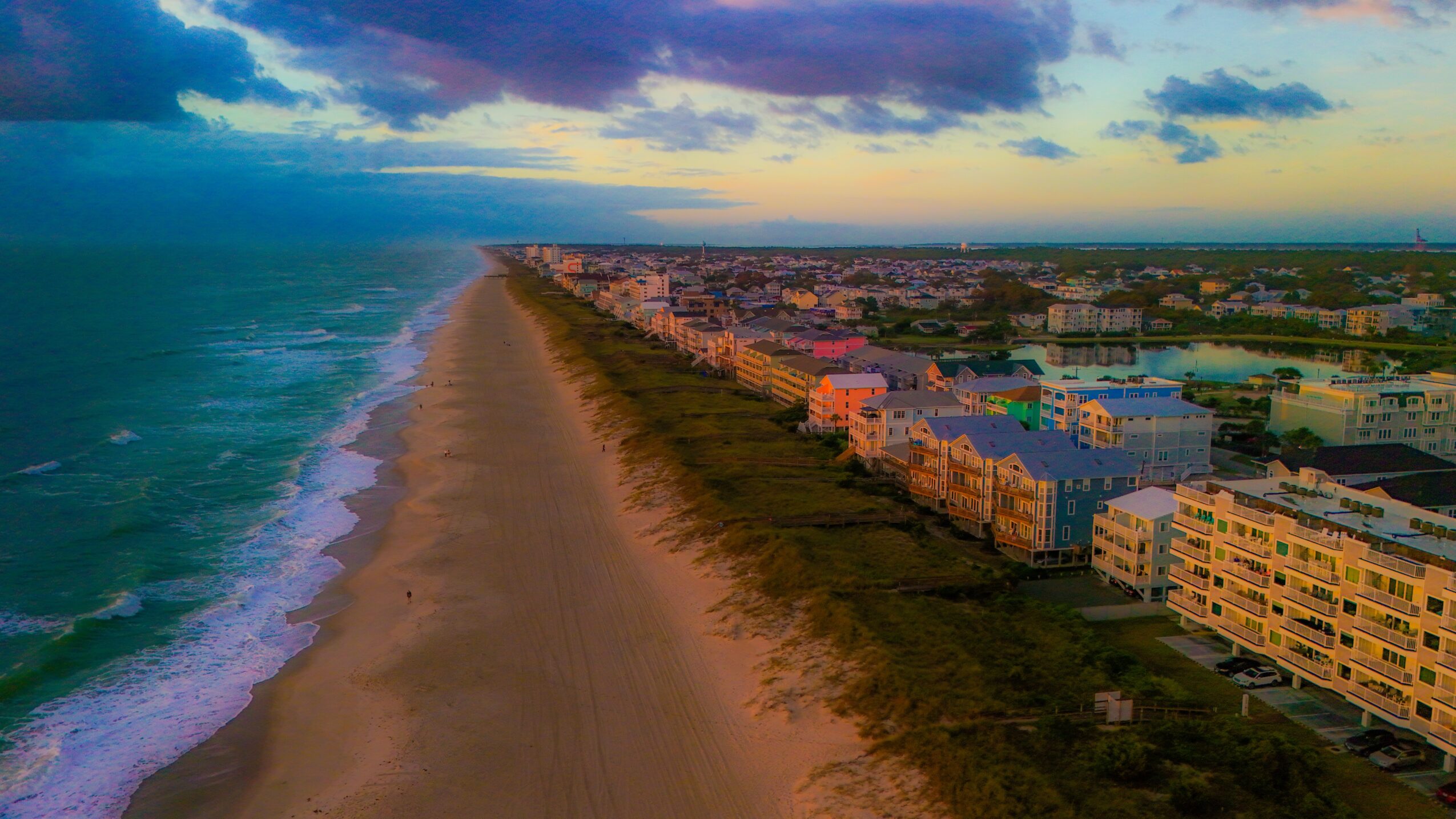 Aerial view of a beach with ocean on the left and buildings on the right, under a partly cloudy sky.