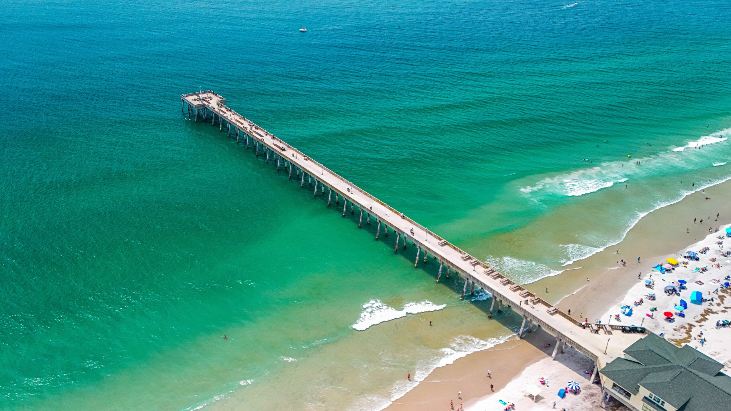 A long pier extends into the ocean with a sandy beach and umbrellas nearby, viewed from above.