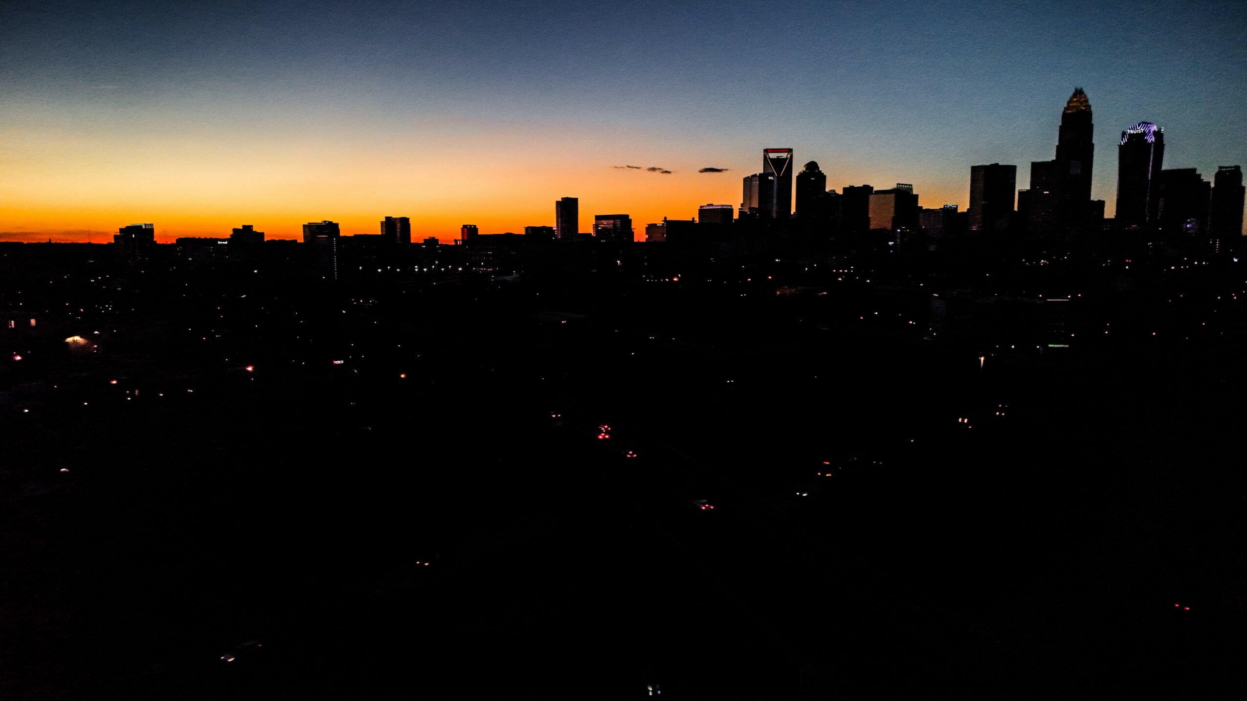 City skyline at sunset with dark buildings and a colorful sky, some lights visible in the cityscape.