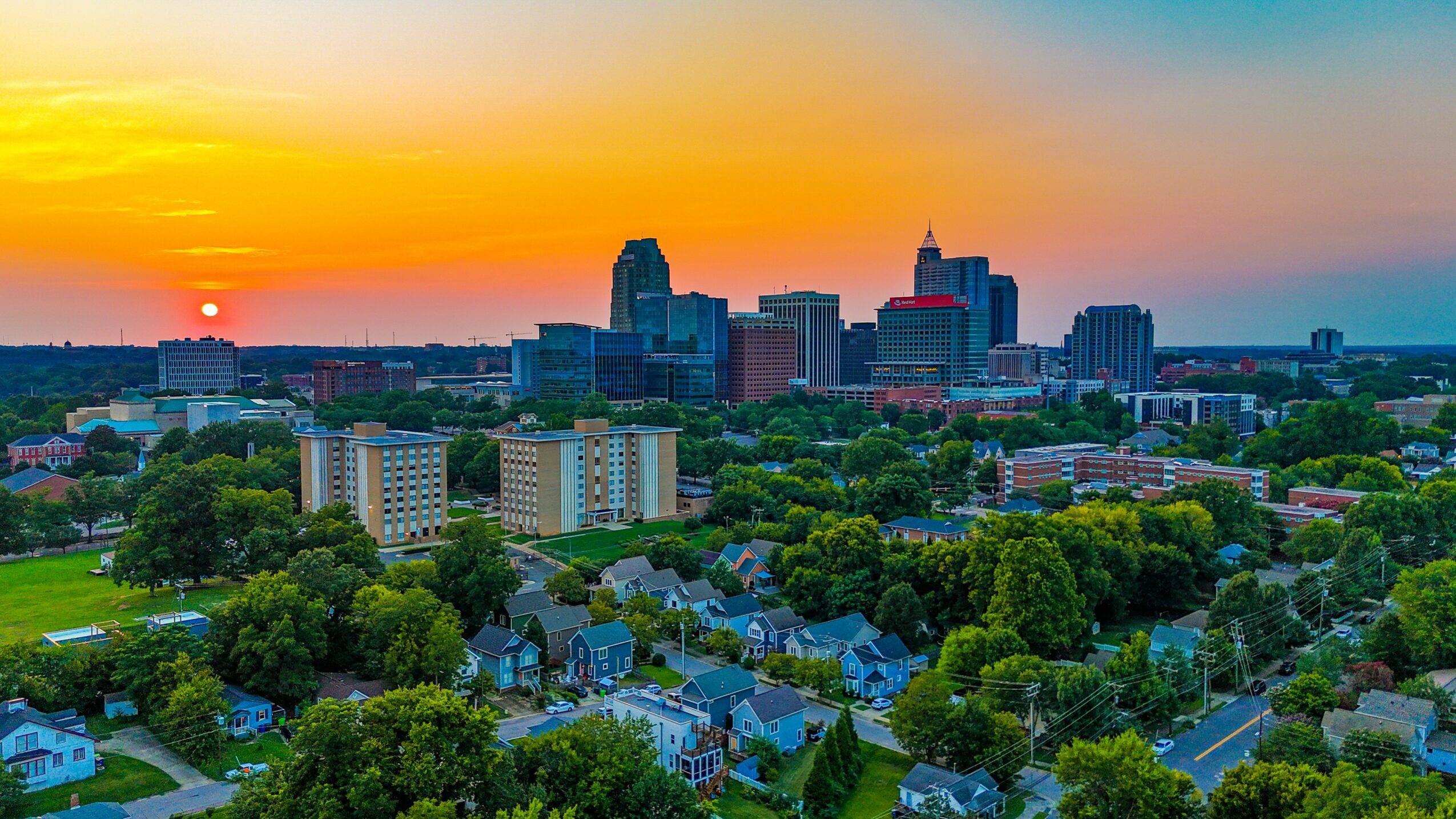 City skyline at sunset with tall buildings, surrounded by green trees and residential areas.