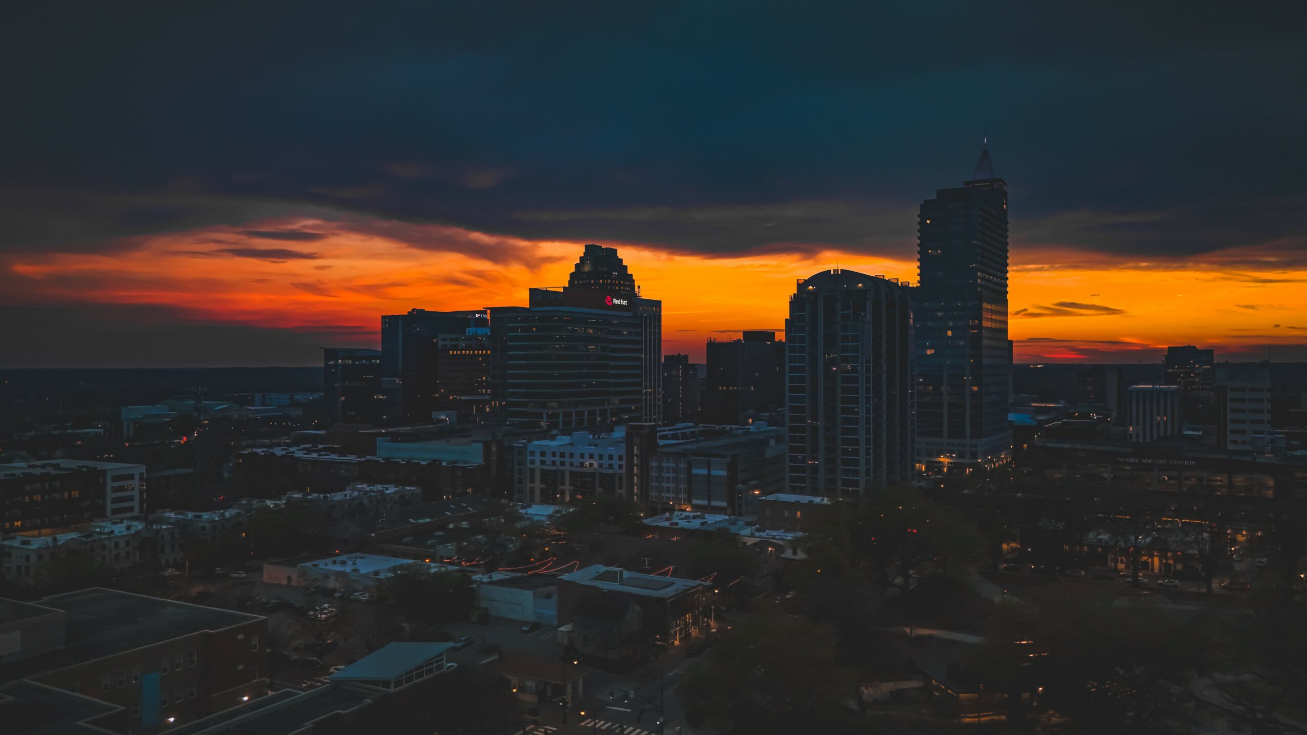 City skyline at sunset with dark clouds and orange sky, silhouetted buildings, and a few lit windows.