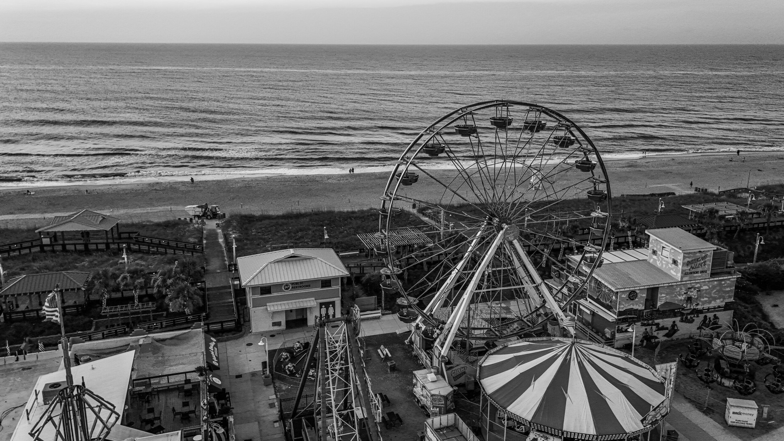 Black and white photo of a seaside amusement park with a Ferris wheel and a striped tent, overlooking the ocean.