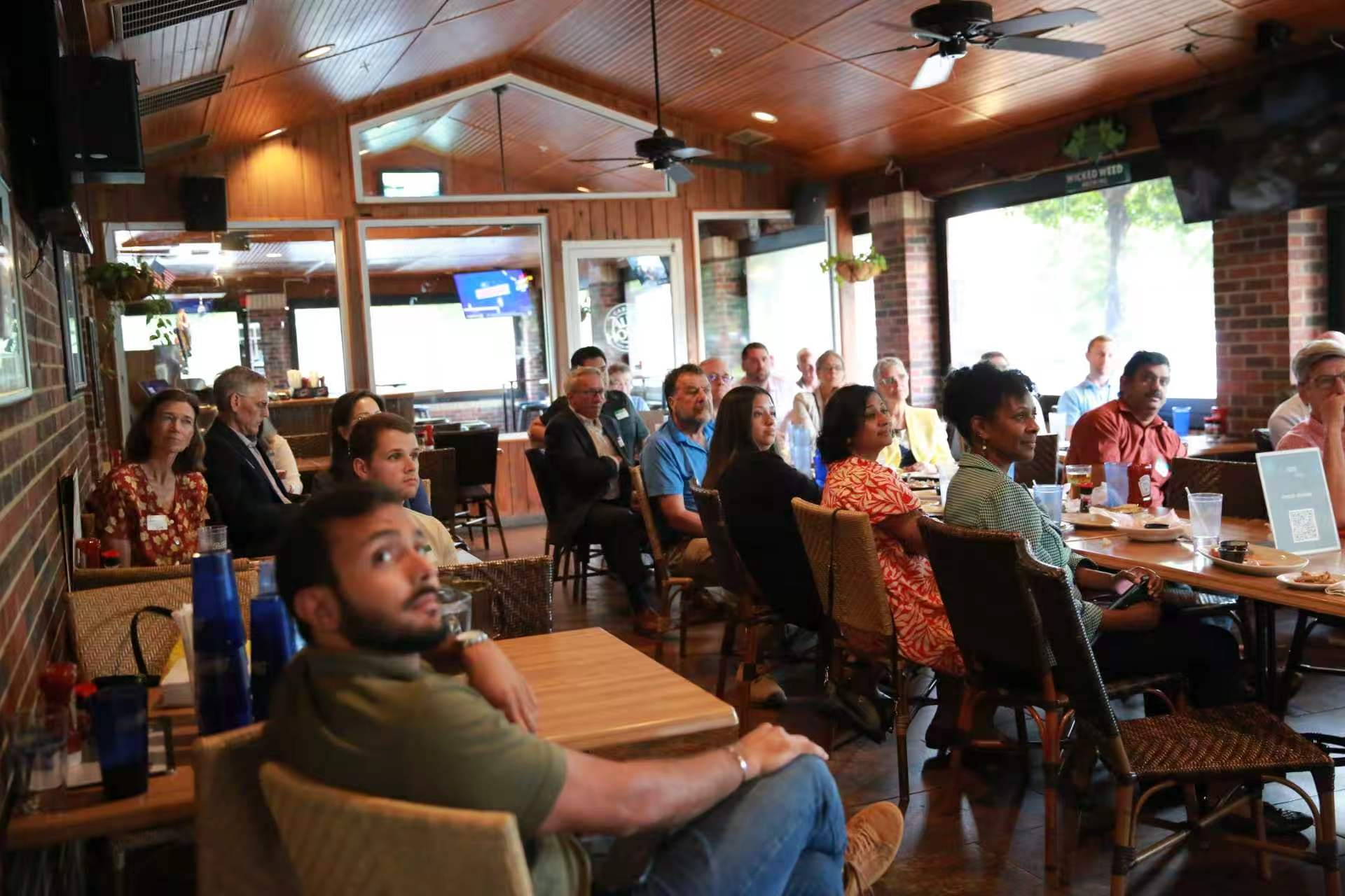 People seated at tables in a restaurant or cafe, facing towards a screen or stage, with a wooden ceiling and brick walls.