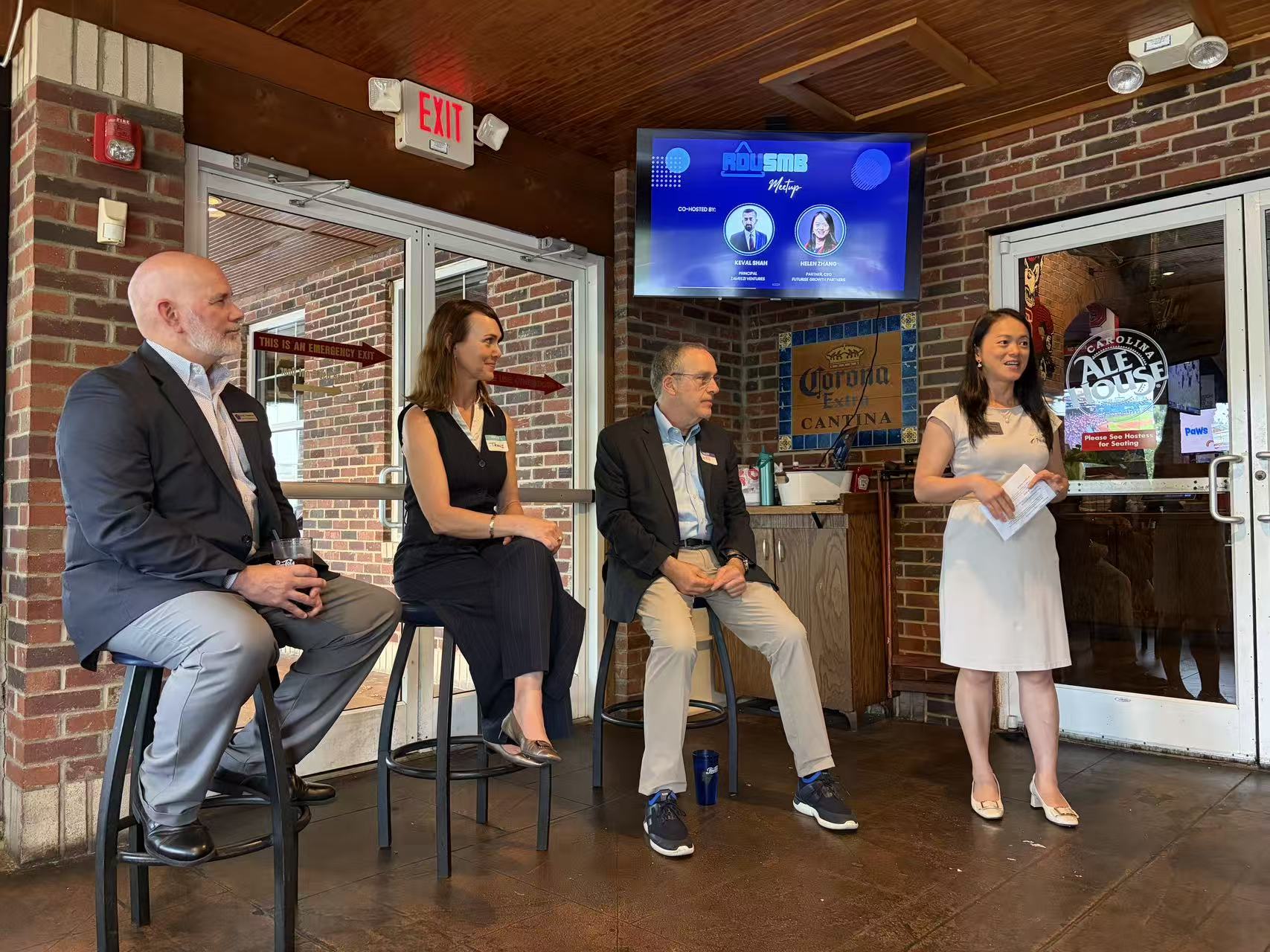 Four people seated and standing in a restaurant or bar, with a brick wall, TV screen, and door behind them.