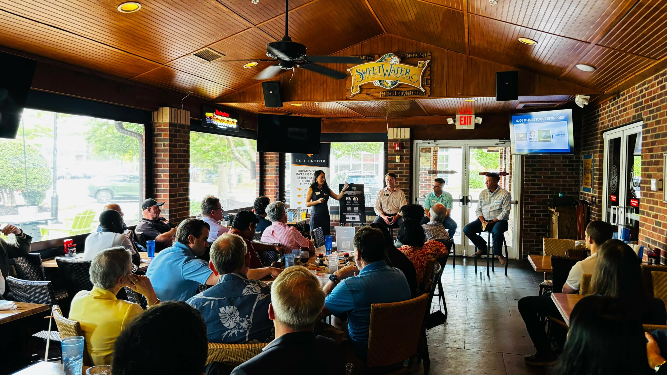 People seated in a restaurant or bar listening to a speaker on stage, with large windows and brick walls.