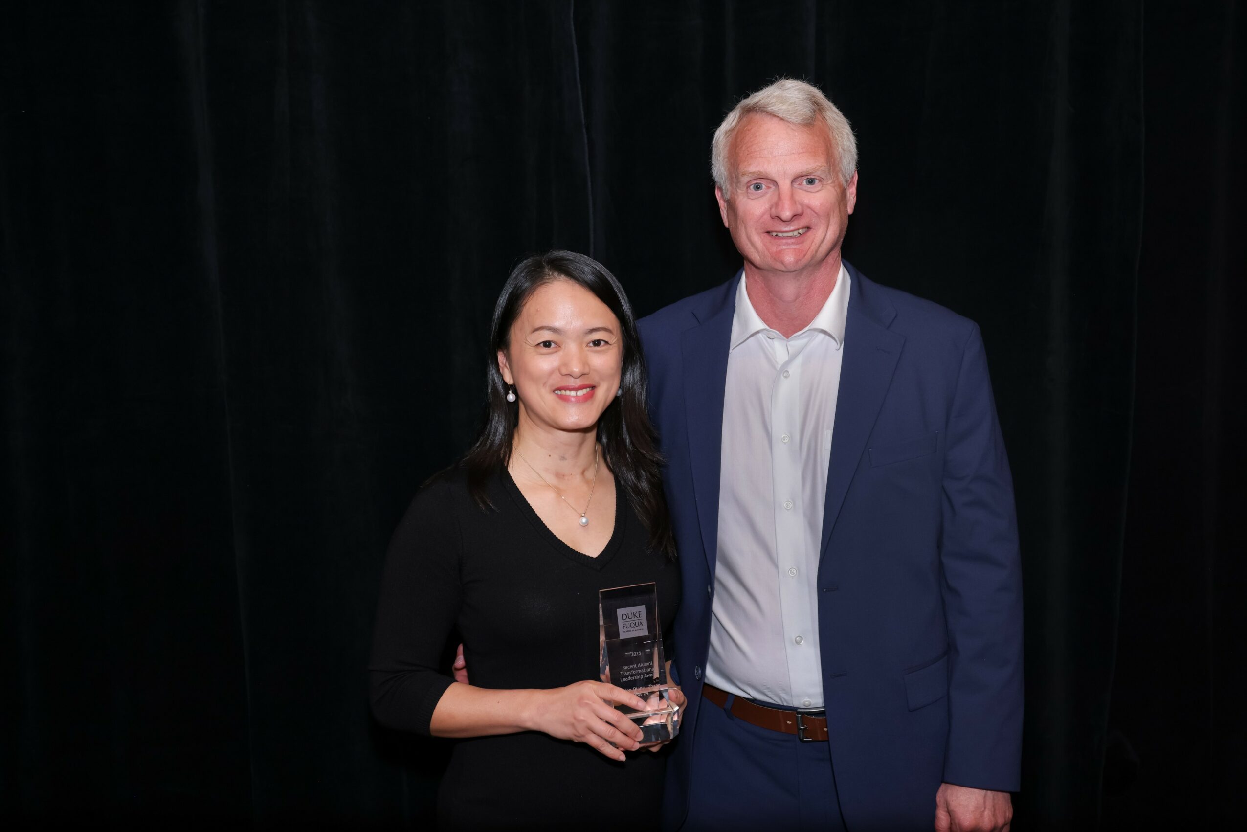 A woman and a man stand together against a black background, smiling, with the woman holding an award or plaque.