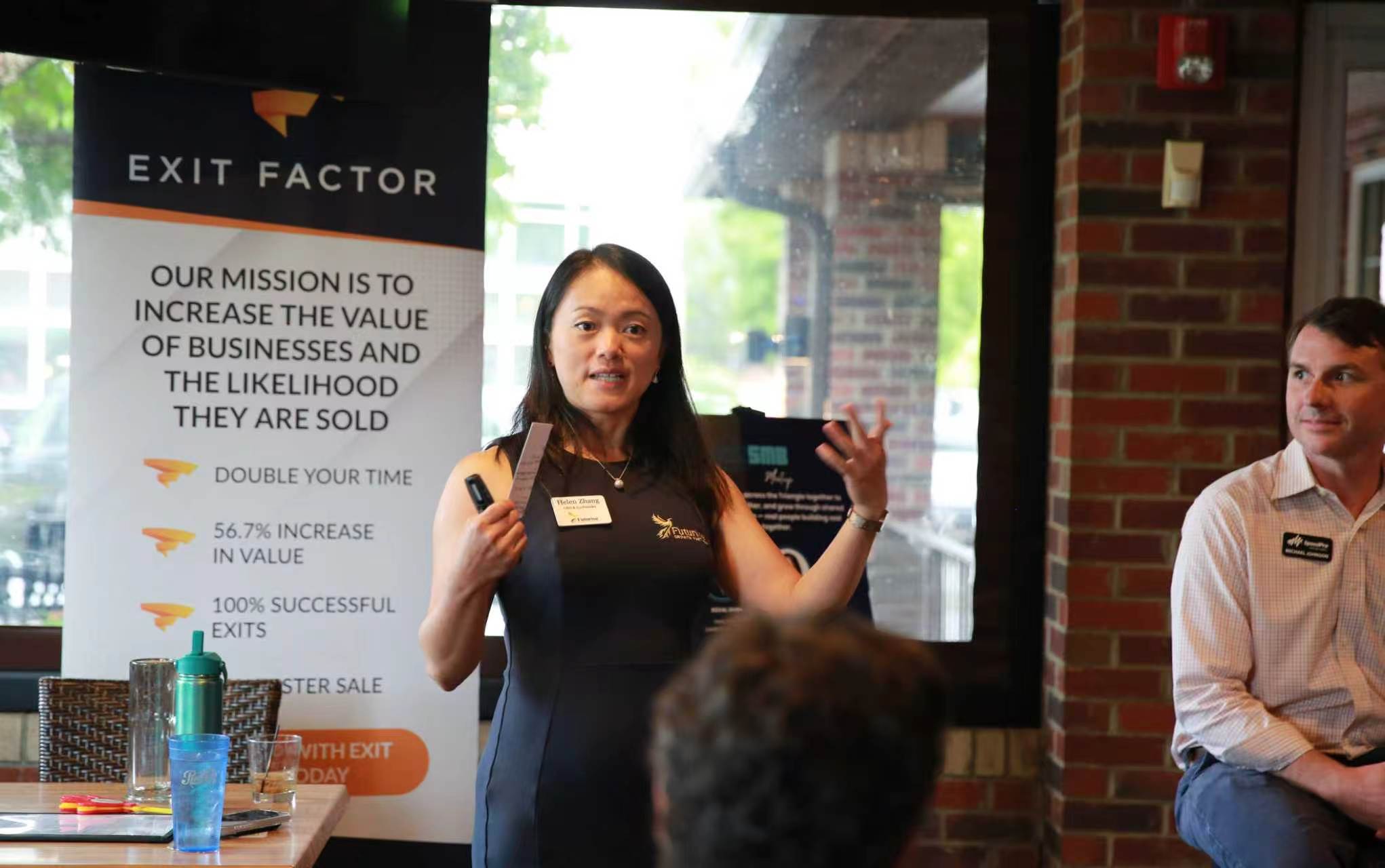 Woman giving a presentation in front of a screen with text, audience members visible, brick wall background.