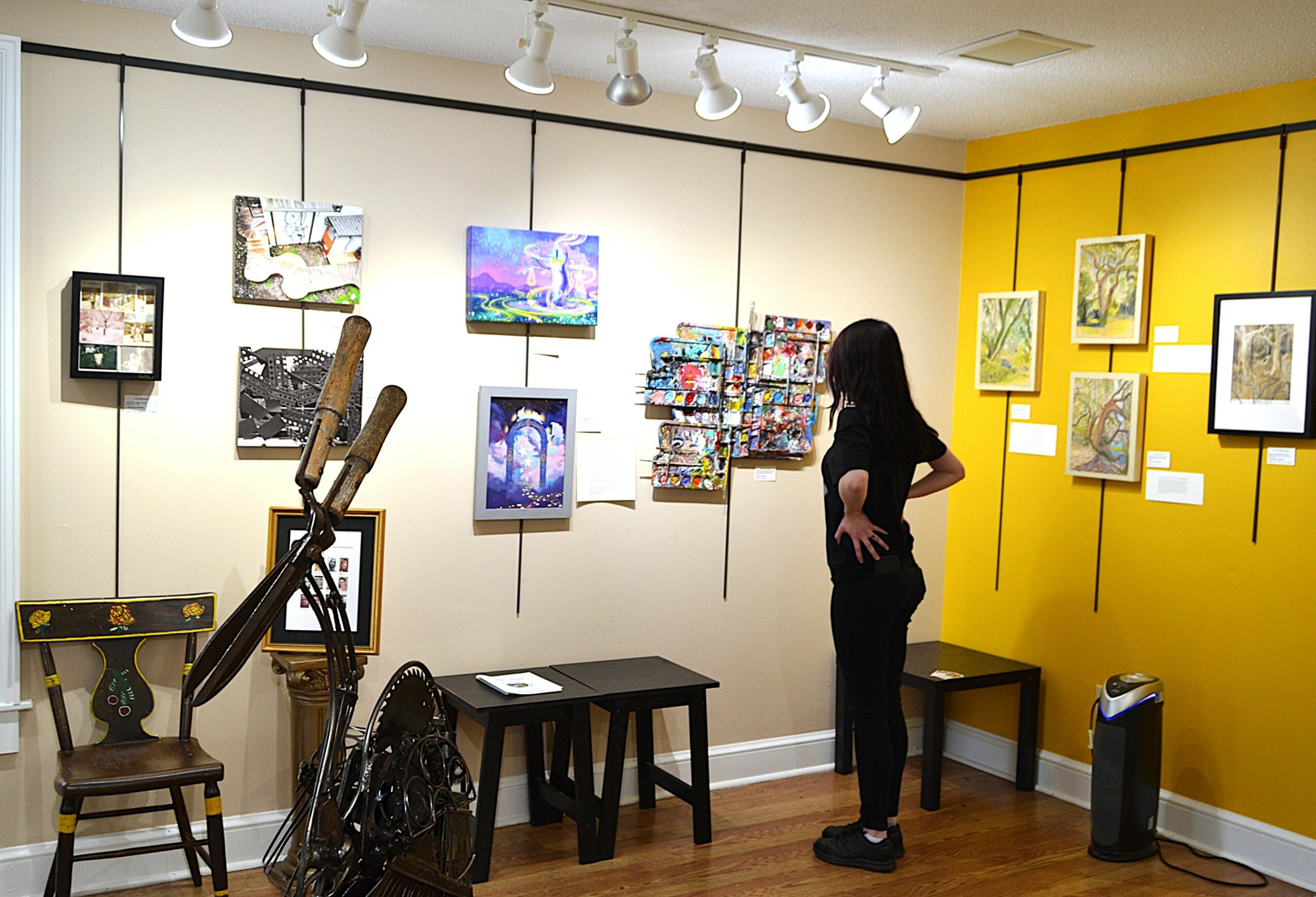 Person viewing artwork in gallery with yellow and white walls, various framed pieces, and a small table.
