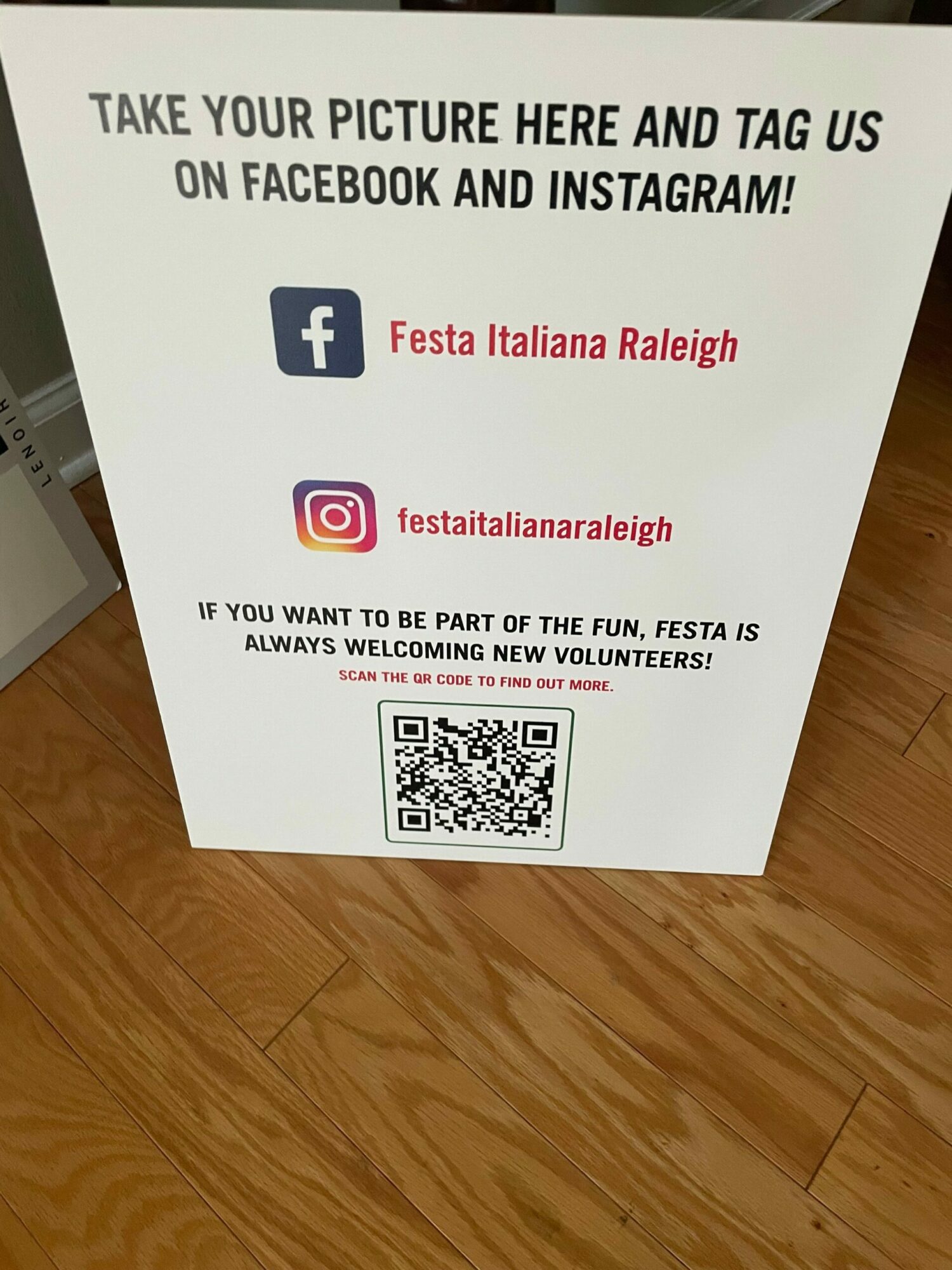 White sign with black and pink text, Facebook and Instagram logos, QR code, on wooden floor.