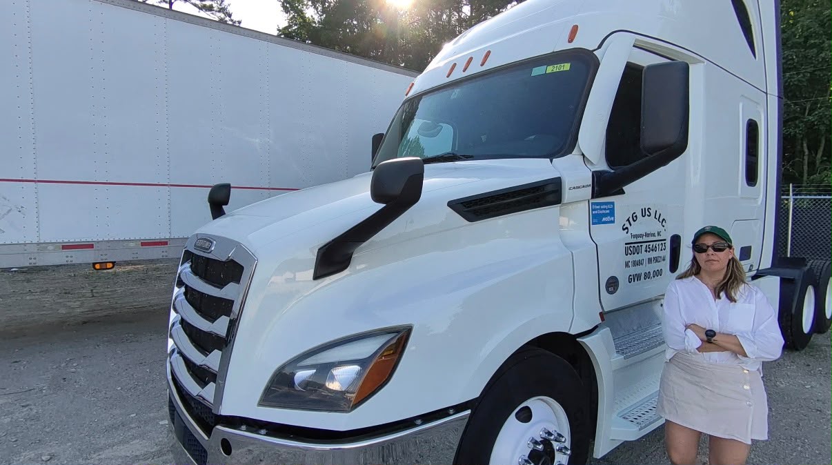 Woman wearing sunglasses and a white dress standing next to a large white semi-truck.