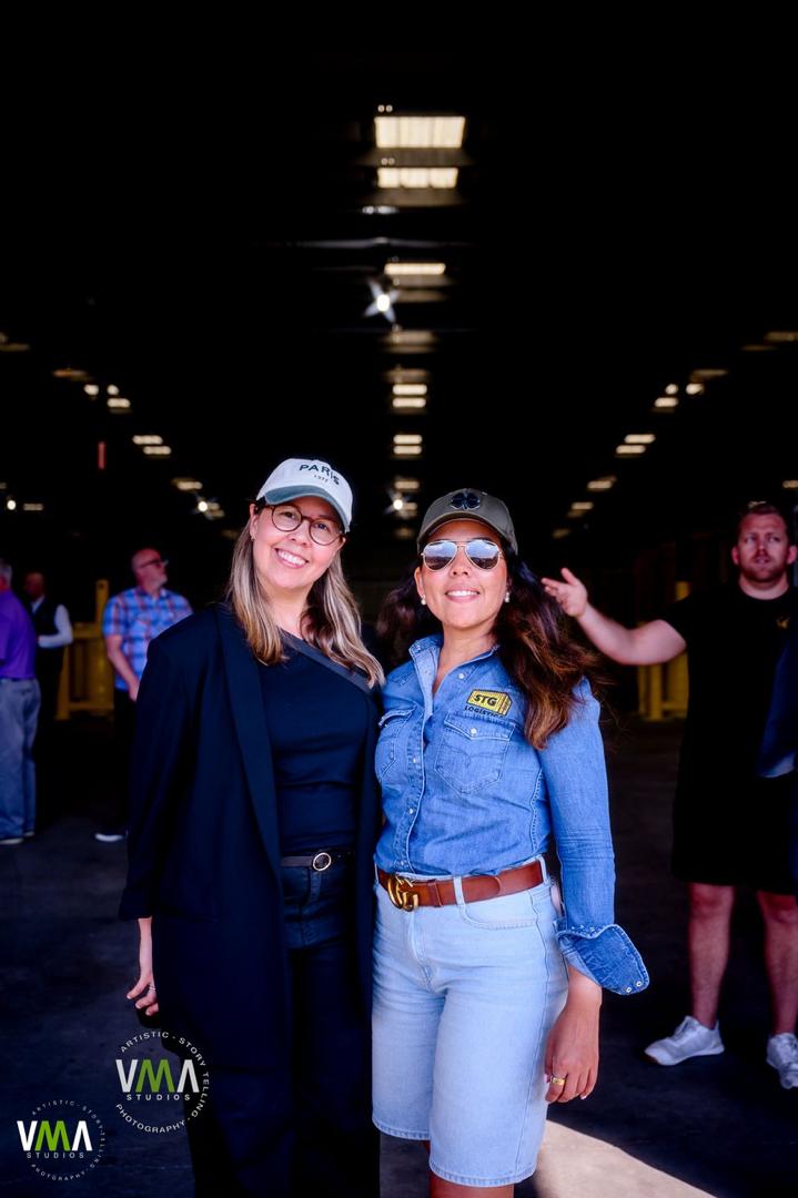 Two women smiling and posing indoors, with people in the background, under ceiling lights, one wearing glasses and a cap.