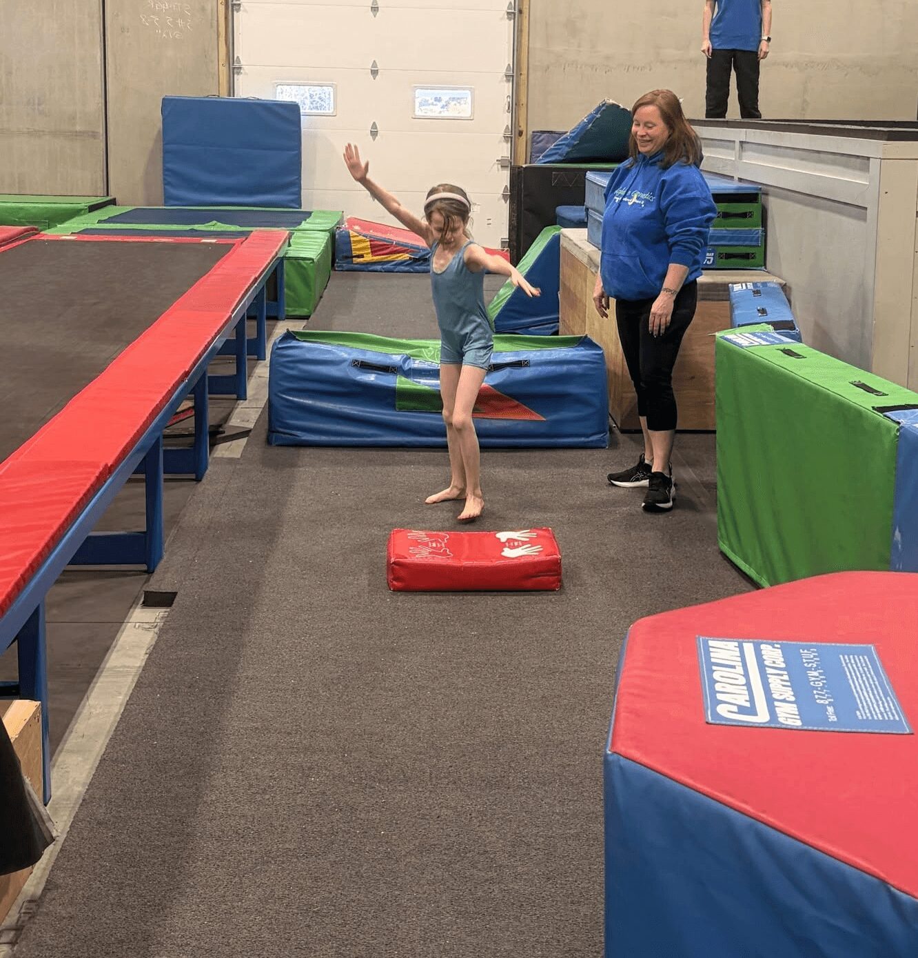 Child jumping on red mat with instructor in an indoor trampoline park, colorful padded equipment around.