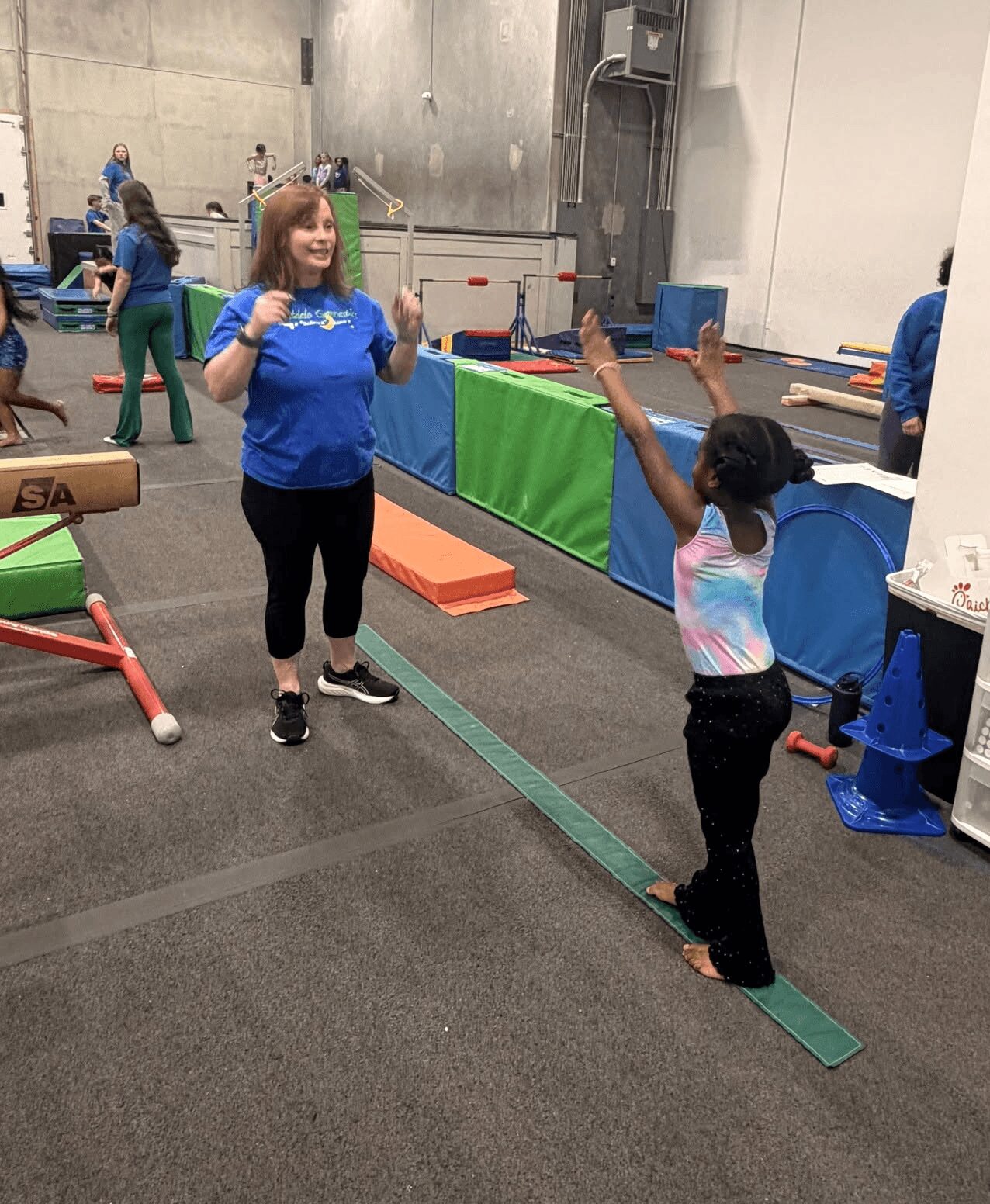 Two children in a gym, one girl with brown hair and a blue shirt, another girl with dark hair and a colorful shirt, standing on a green balance beam, smiling and talking.