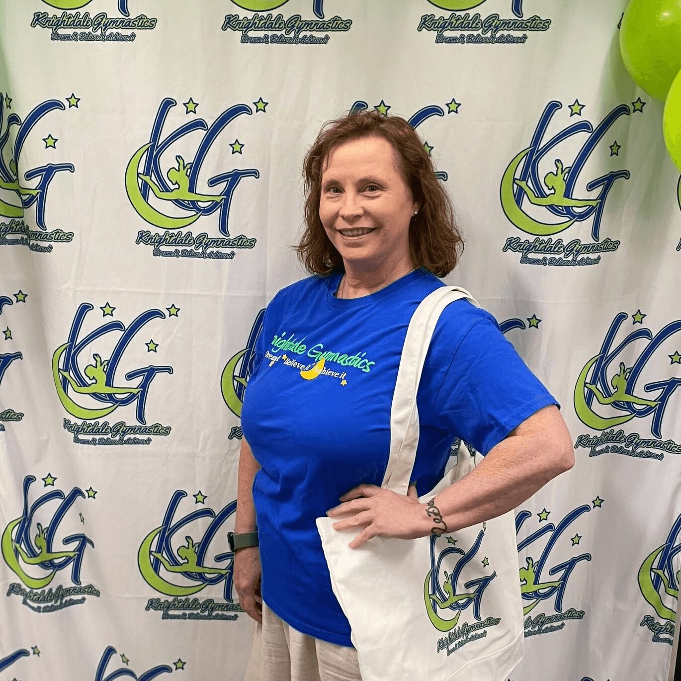 Woman smiling in blue T-shirt standing in front of a backdrop with logos, holding a white tote bag, surrounded by green balloons.