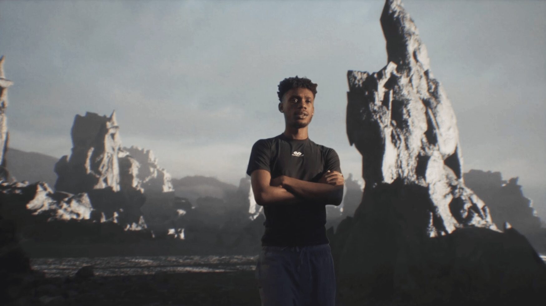 Young man standing with arms crossed outdoors, rocky formations in background, cloudy sky, casual clothing.