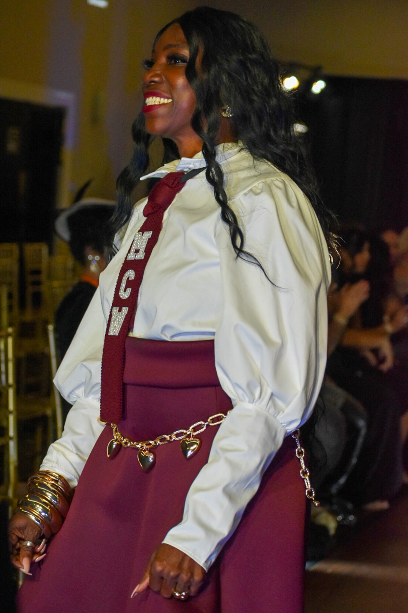 Woman in graduation gown and sash smiling, with long dark hair, in an indoor setting with people in background.