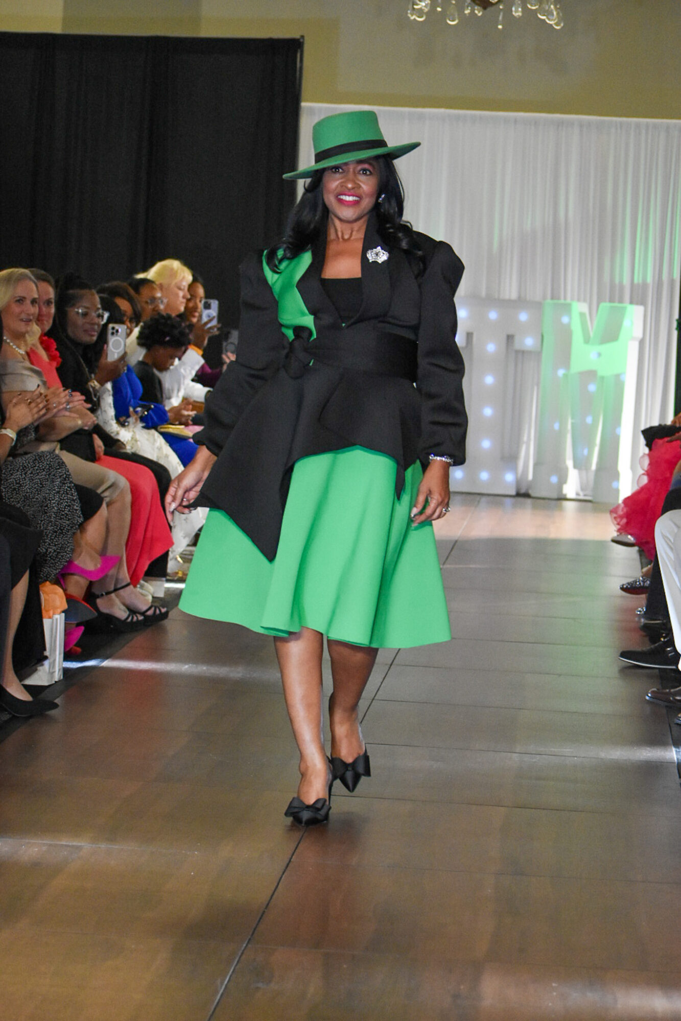 Woman walking on runway wearing a black coat, green dress, green hat, and black heels, with audience watching.