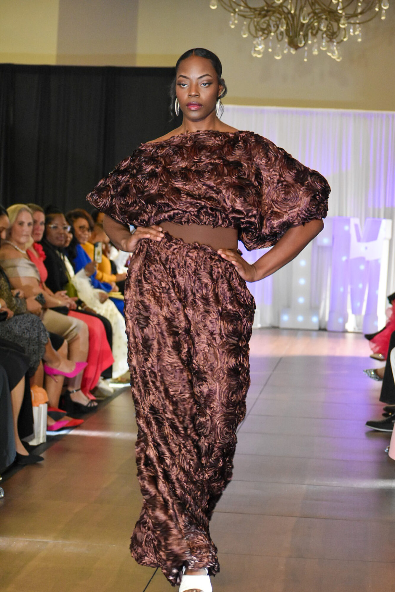 Model walking down runway in brown textured outfit, audience seated on both sides, chandelier overhead.