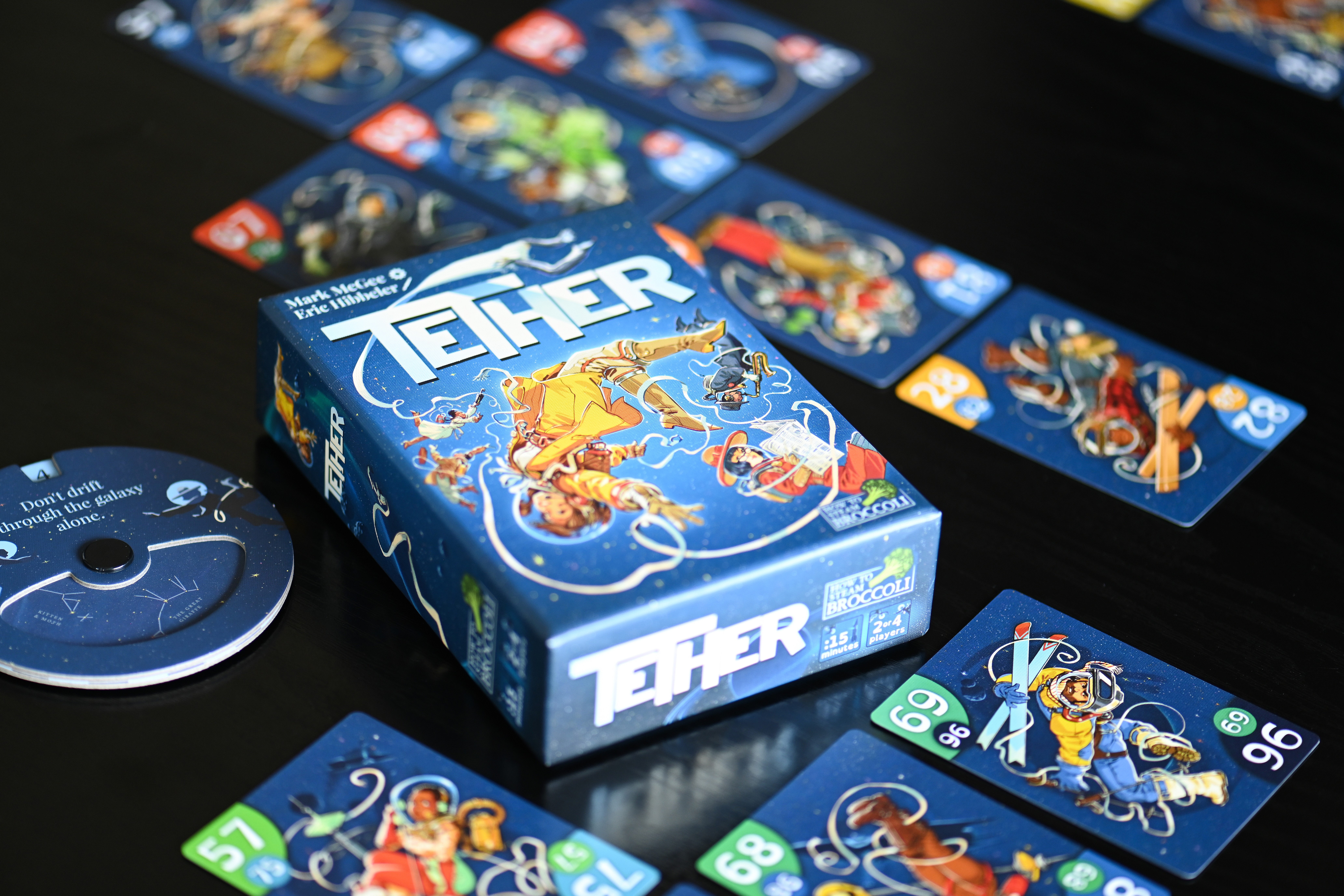 Card game box with colorful illustrations, game cards spread out on a black surface, and a circular game piece nearby.
