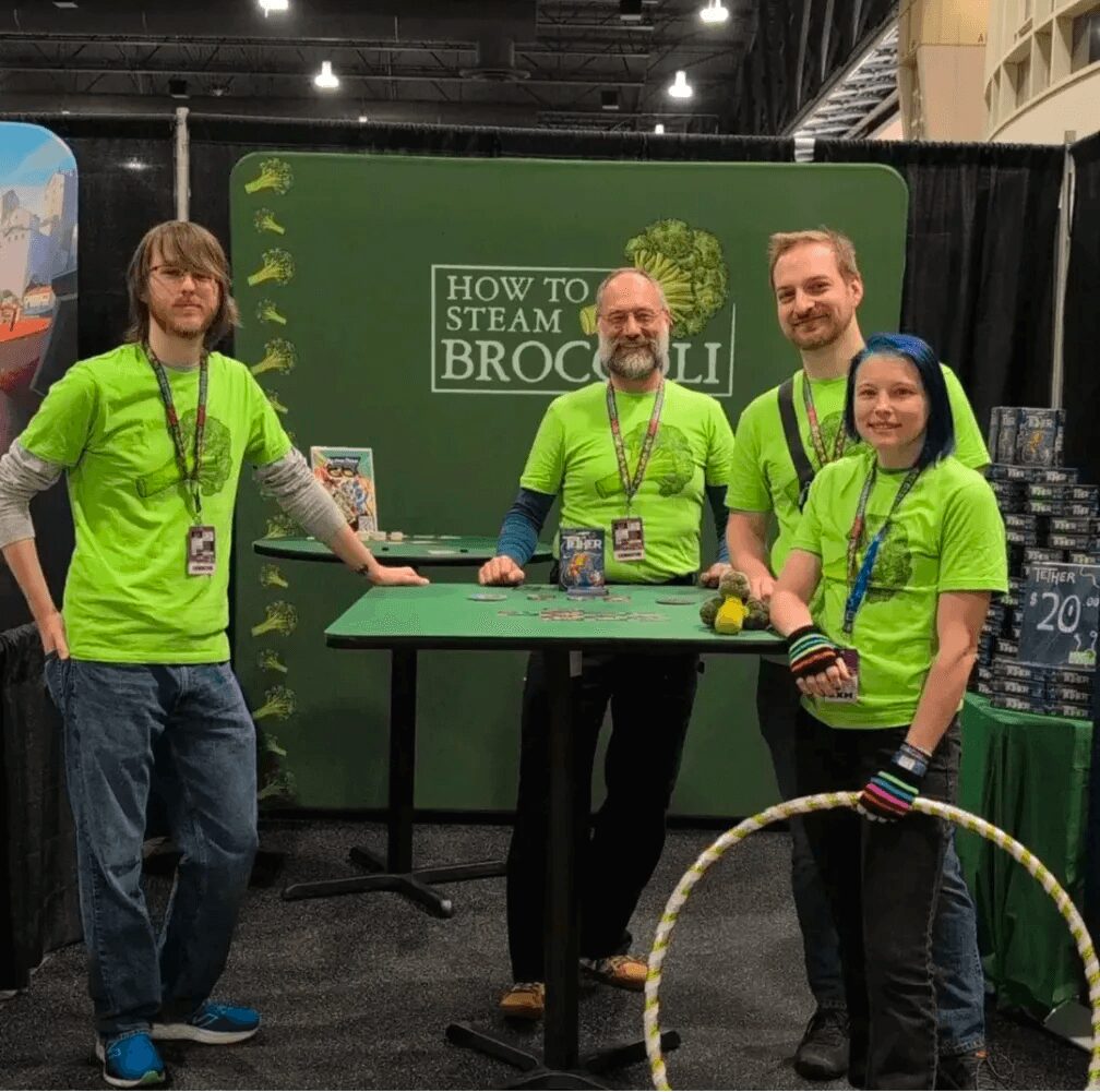 Four people in bright green shirts stand behind a table at a booth with a green backdrop reading 'How to Steam Broccoli.'