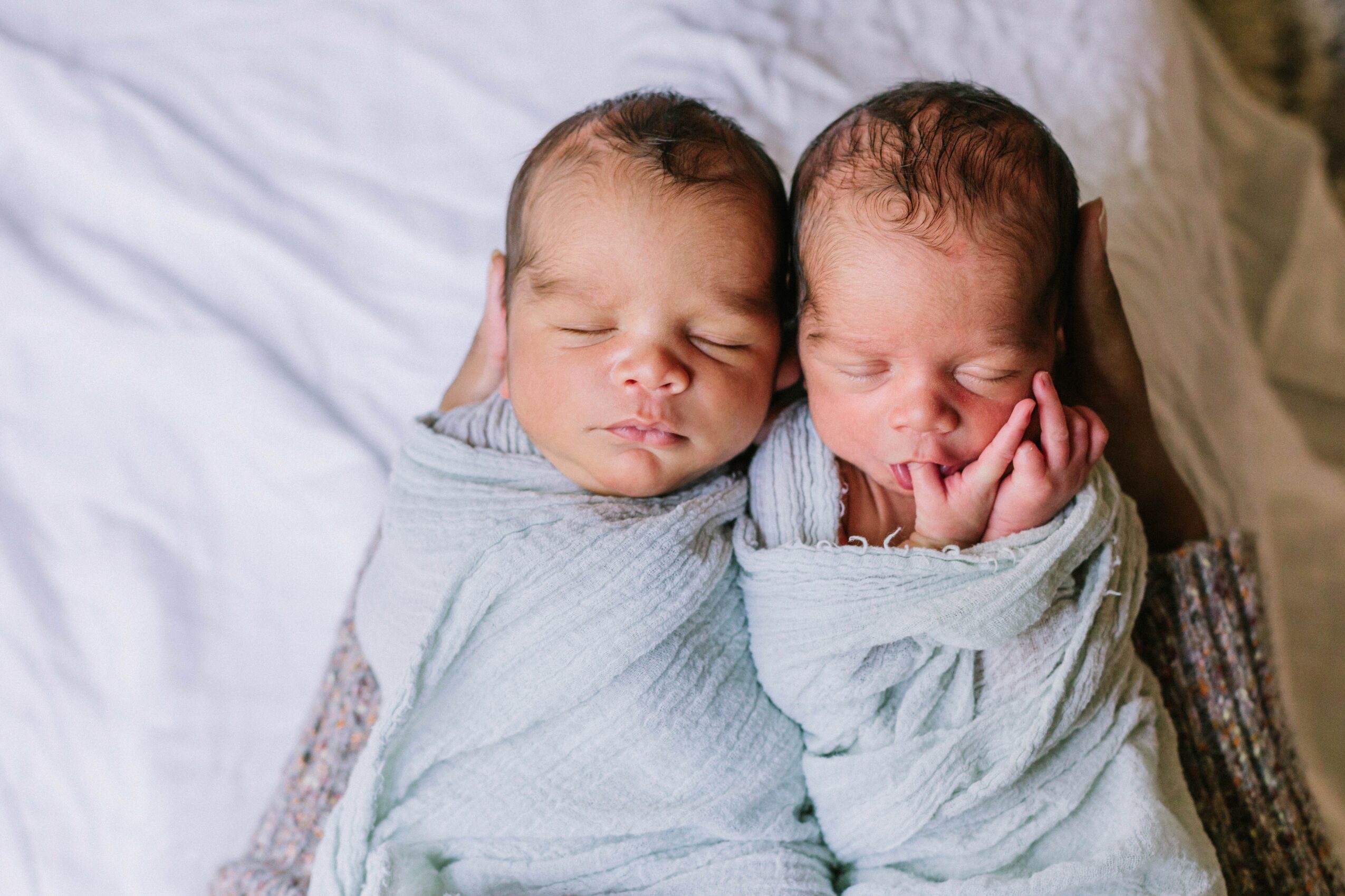 Two newborn babies wrapped in blankets, sleeping side by side on a bed.
