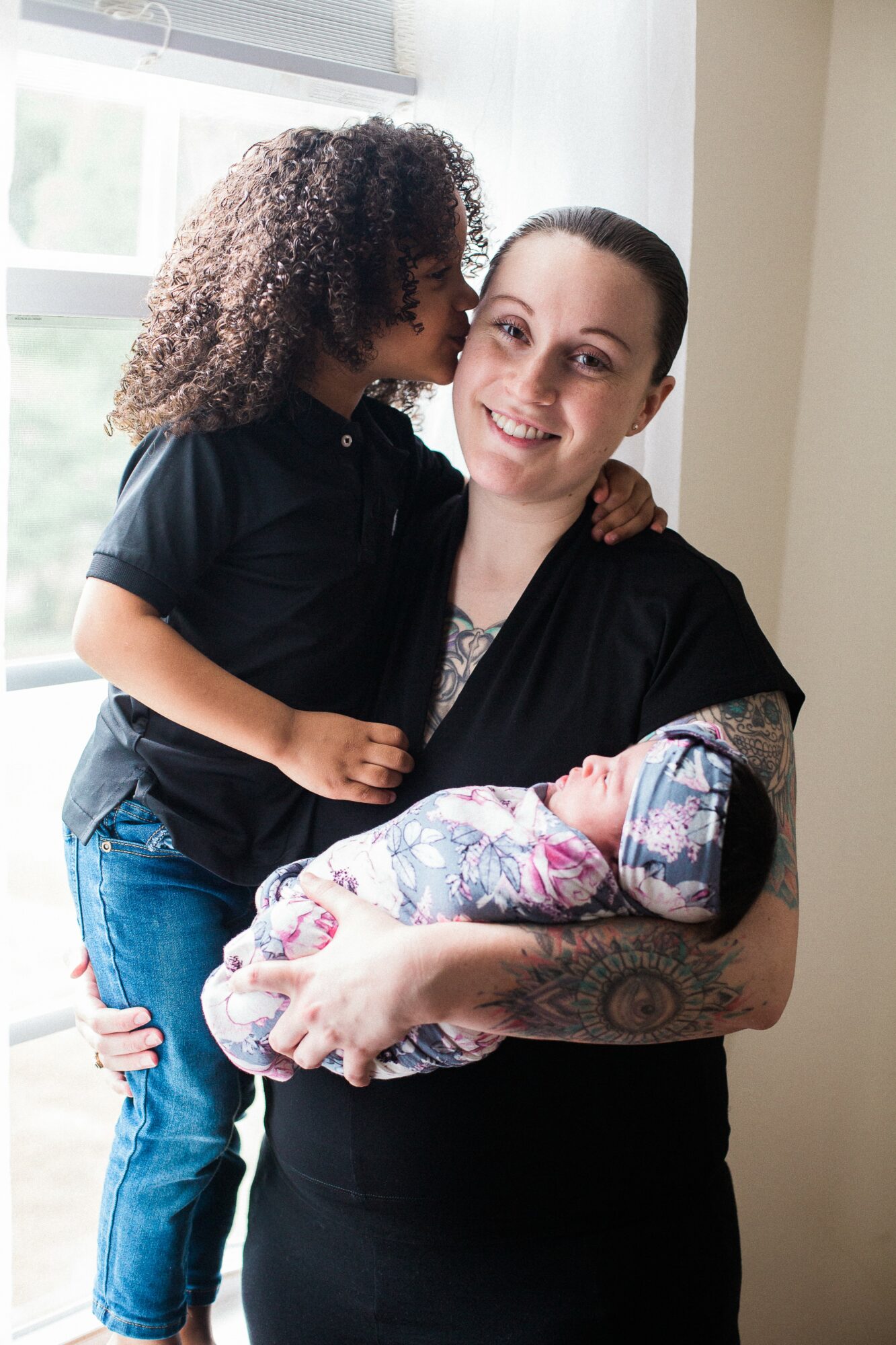 Woman holding a newborn baby, with a young girl kissing her cheek, indoors near a window.