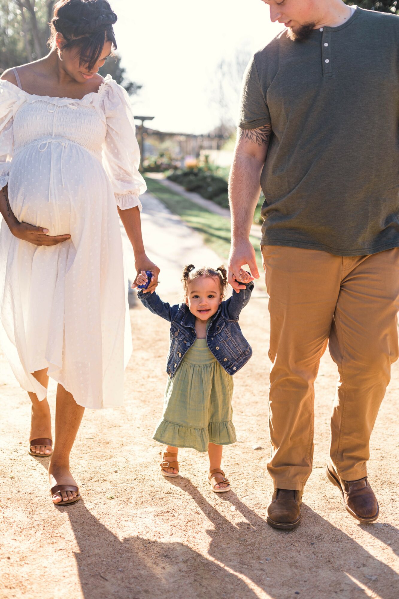 A family of three walking outdoors, holding hands, with a young girl in the center, sunlight shining, and trees in background.