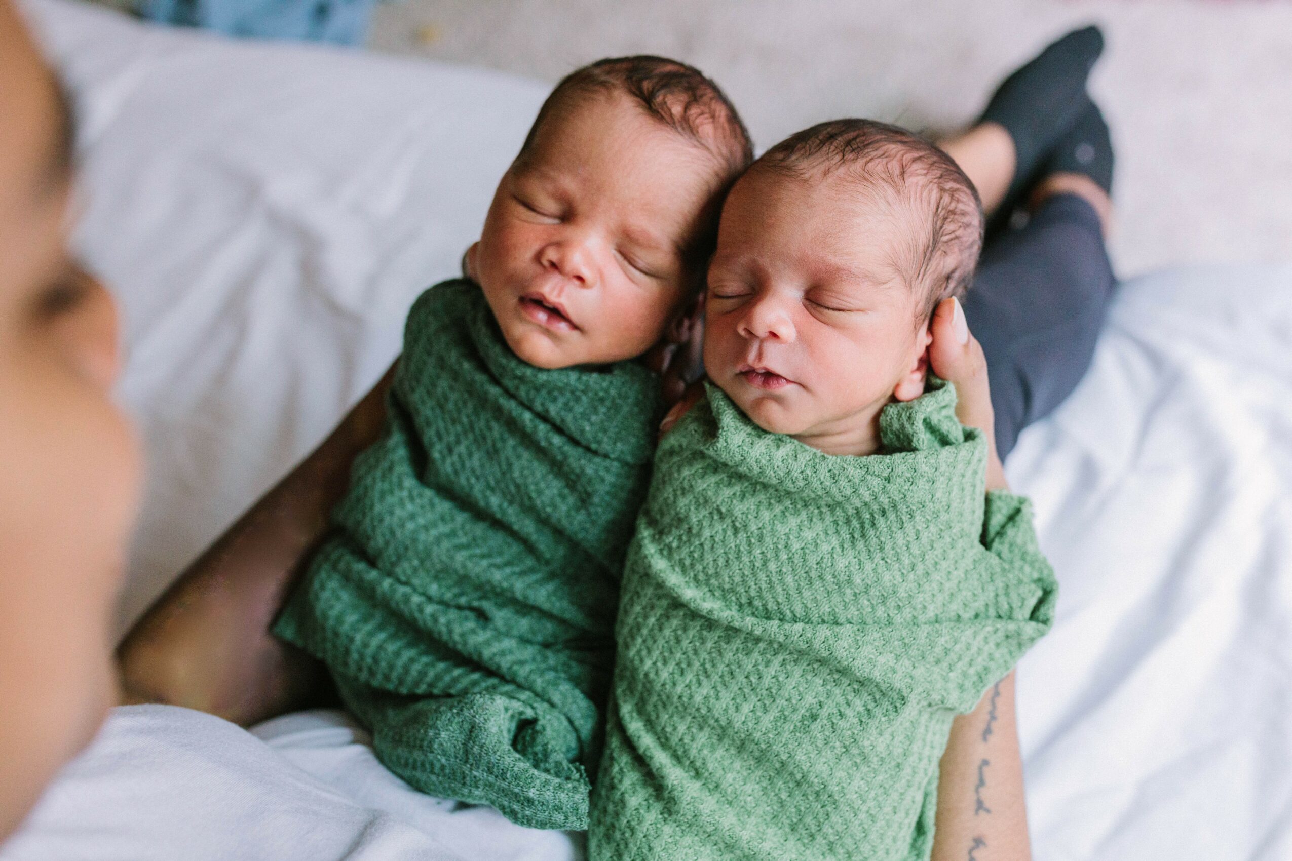 Two newborn babies wrapped in green blankets lying close together on a bed, with a person partially visible nearby.