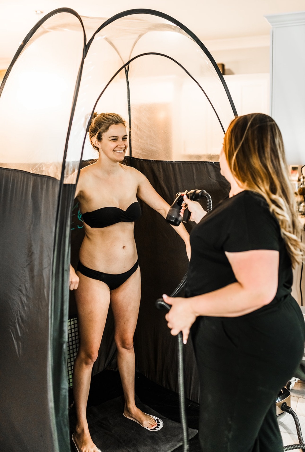 Woman in black bikini being photographed inside a tent by a photographer, both smiling, indoor setting.