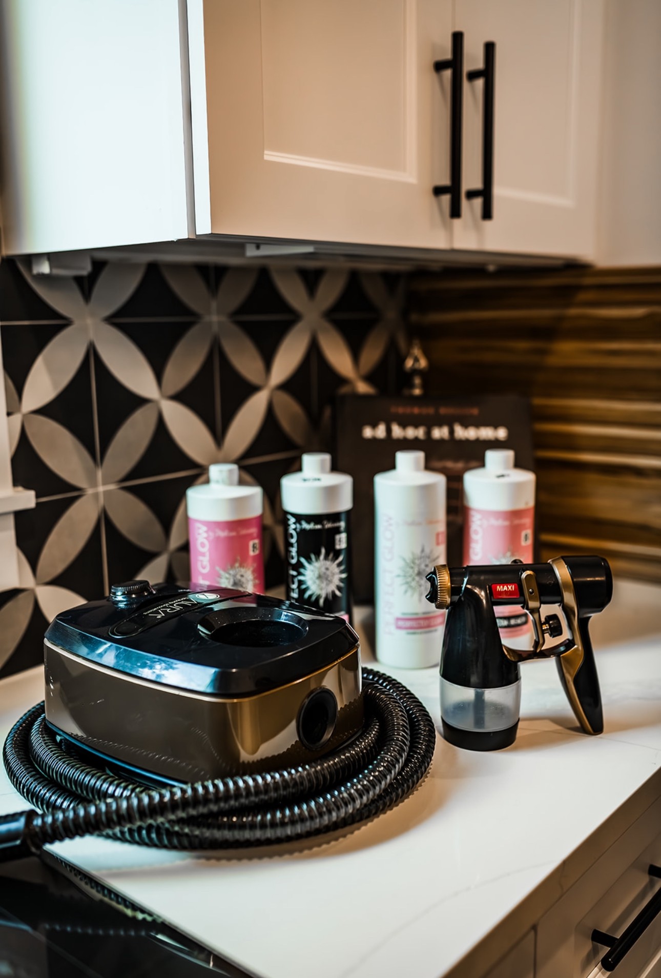 Kitchen countertop with a black appliance, three white bottles, a small black and white device, and a camera, against a patterned backsplash.