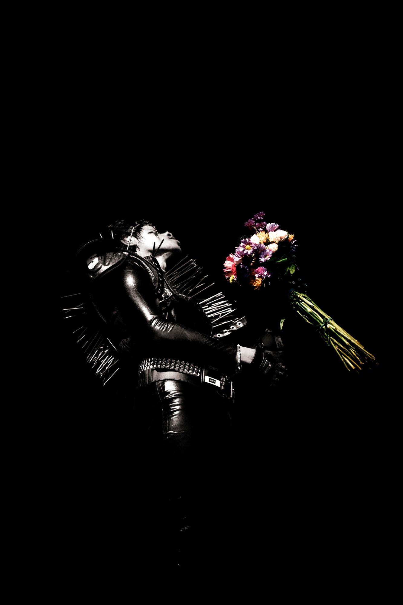 Person in black leather outfit holding a bouquet of colorful flowers against a dark background.