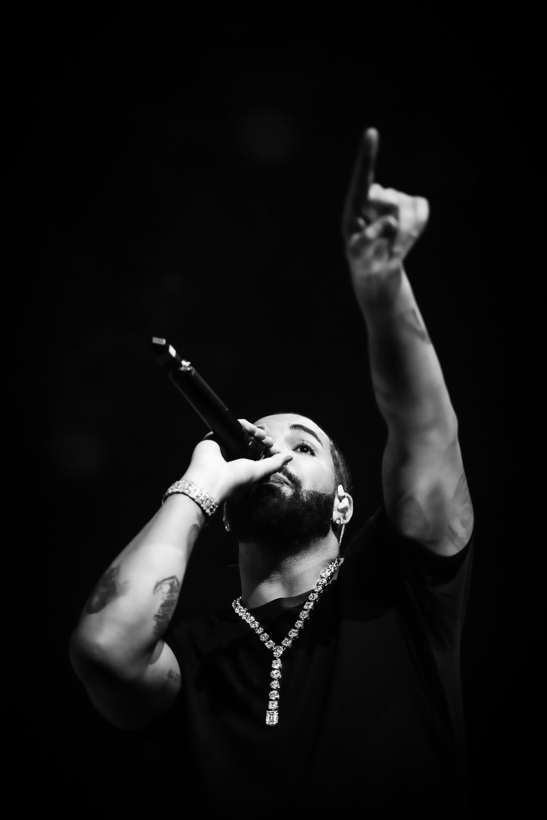 Man with a beard holding a microphone, raising one arm with a finger pointing upward, wearing a necklace, in black and white.