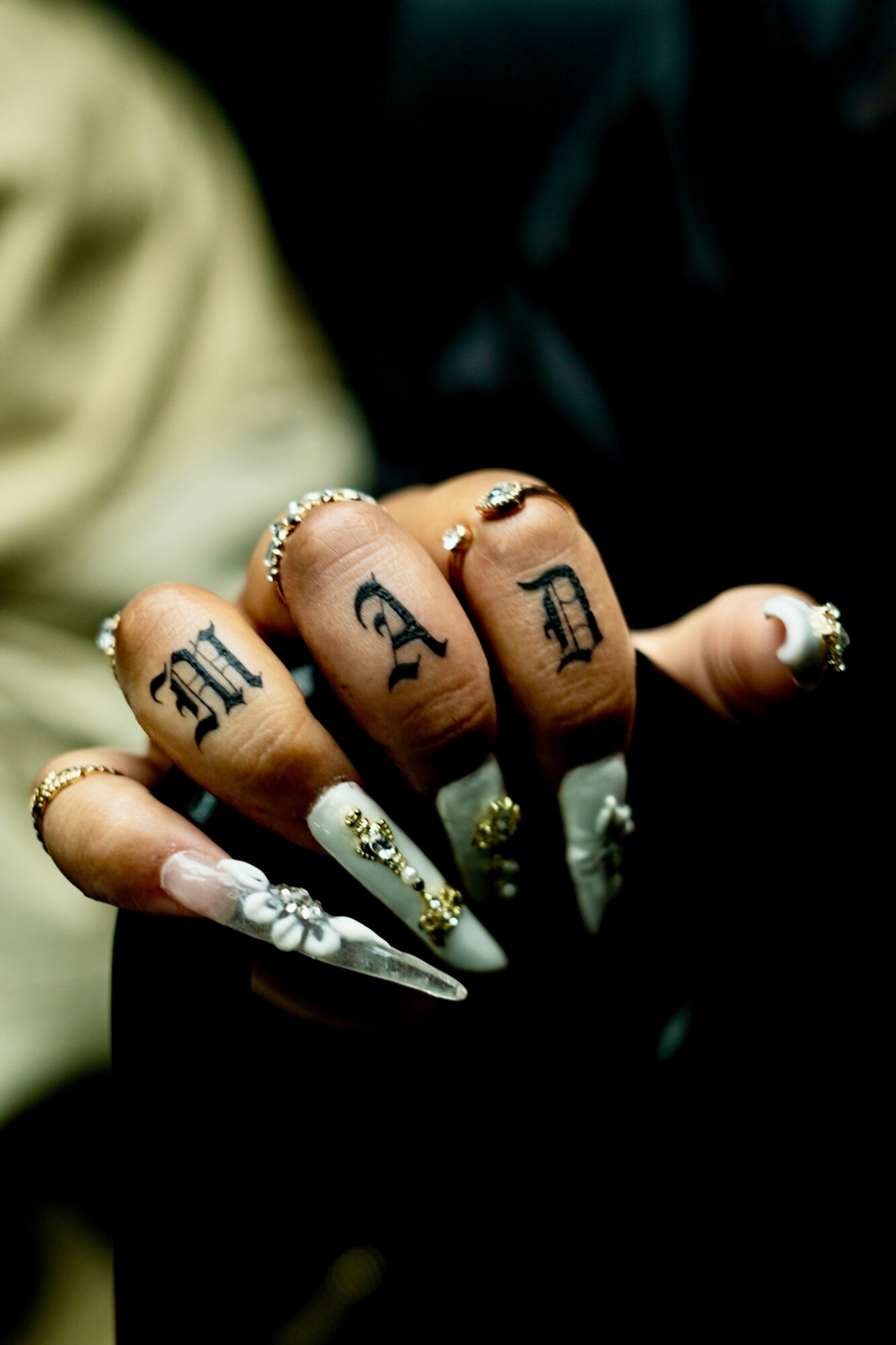 Close-up of a hand with tattoos on fingers spelling 'M A F I A', decorated nails, rings, and long acrylic nails with white floral designs.