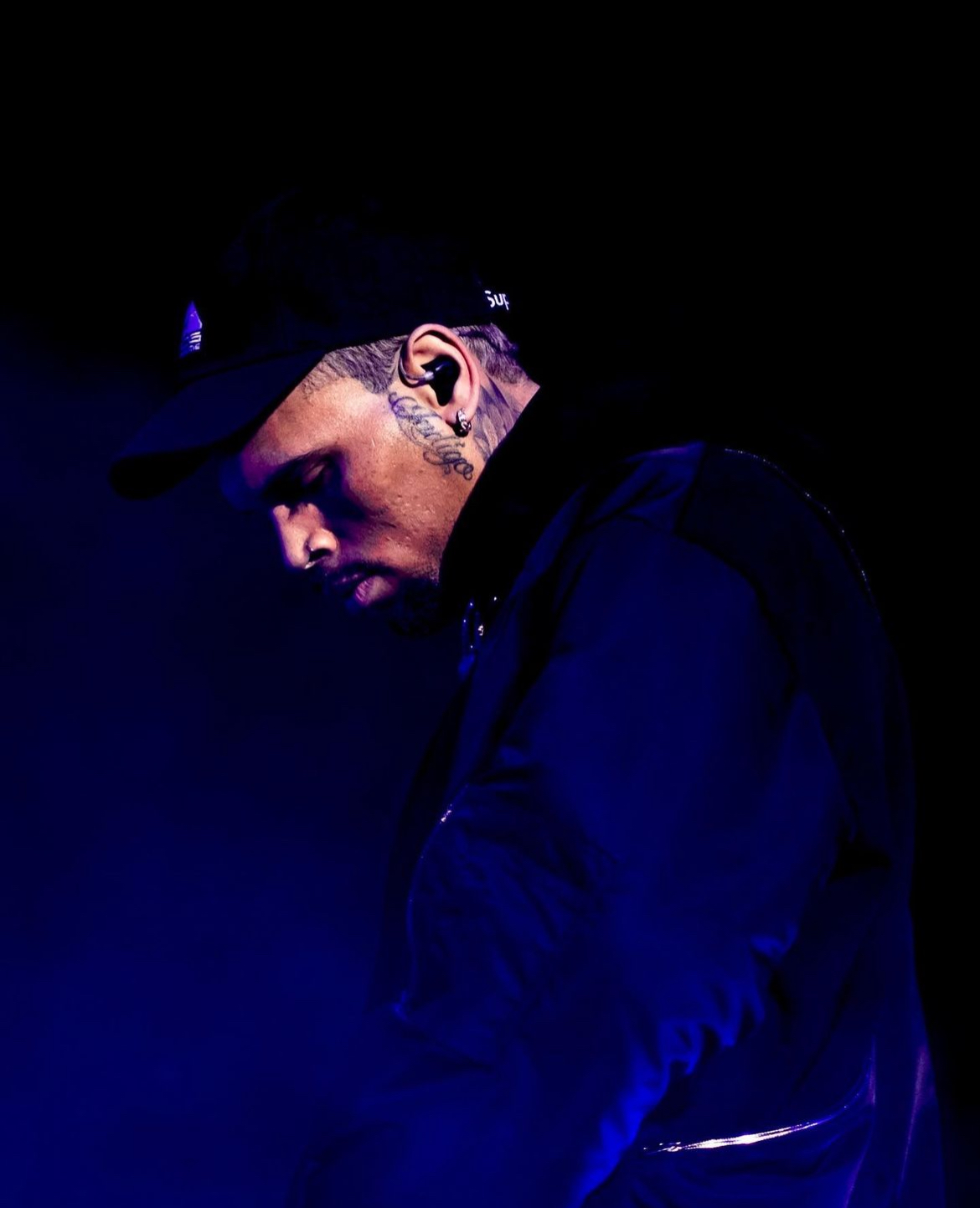 Person with tattoos and earrings, wearing a cap and dark clothing, looking downward in a dark setting.