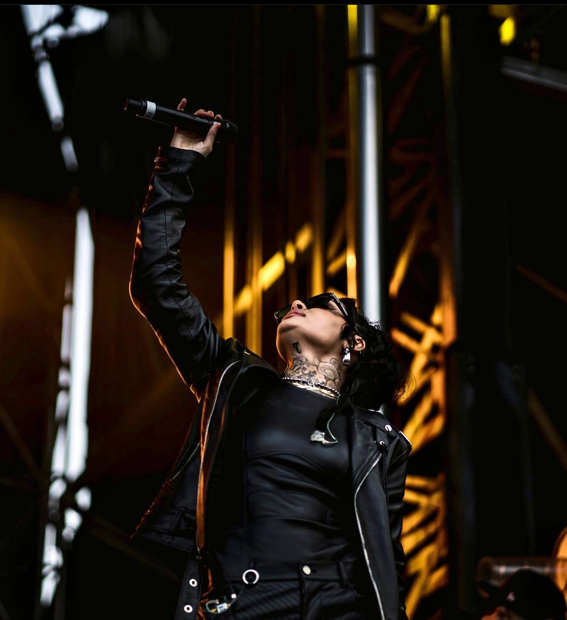 Performer on stage holding microphone, looking upward, with stage lights and structure in background.