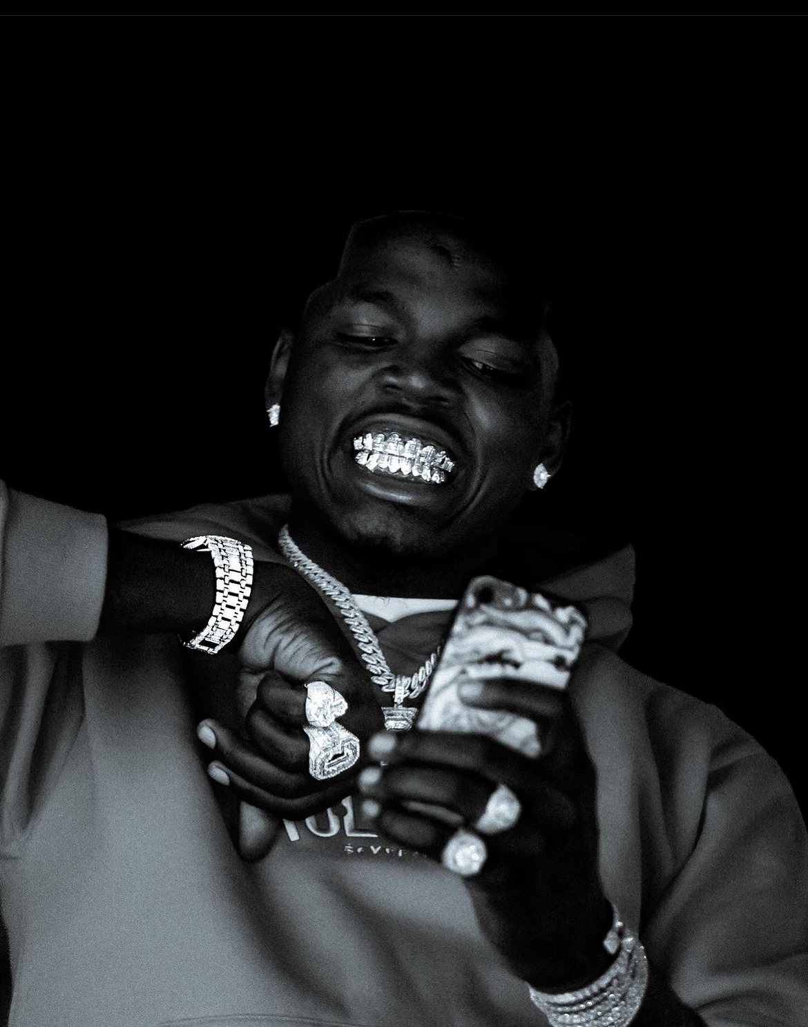 Person with jewelry and grillz looking at a phone, smiling, in a dark background.