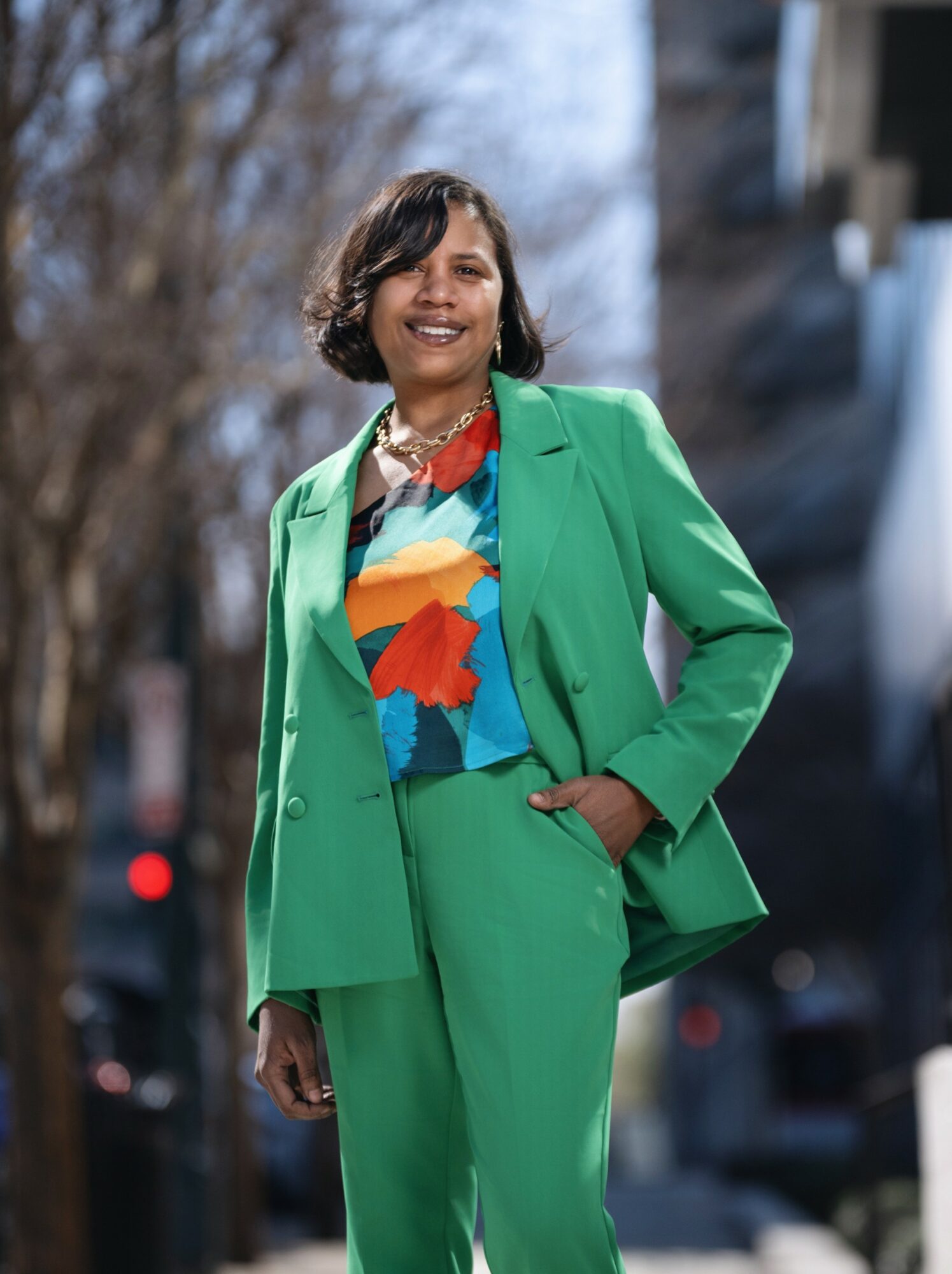 Woman in a bright green suit and colorful blouse standing outdoors, smiling, with trees and buildings in background.