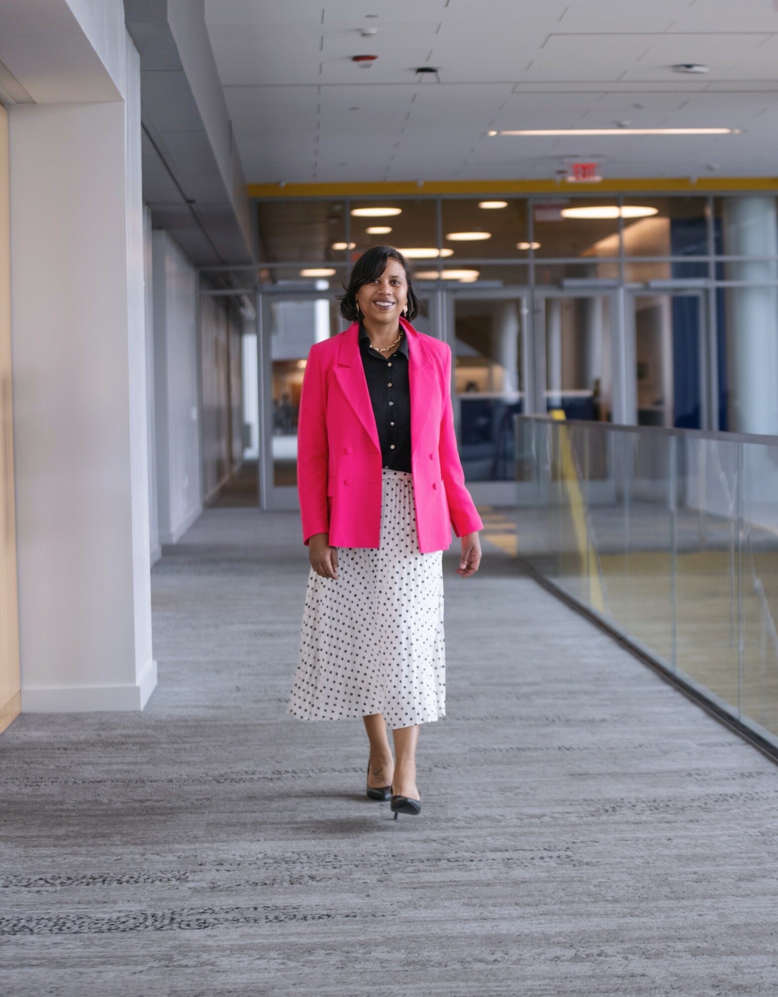 Woman walking in a modern indoor space, wearing a pink blazer and a white skirt with black dots.