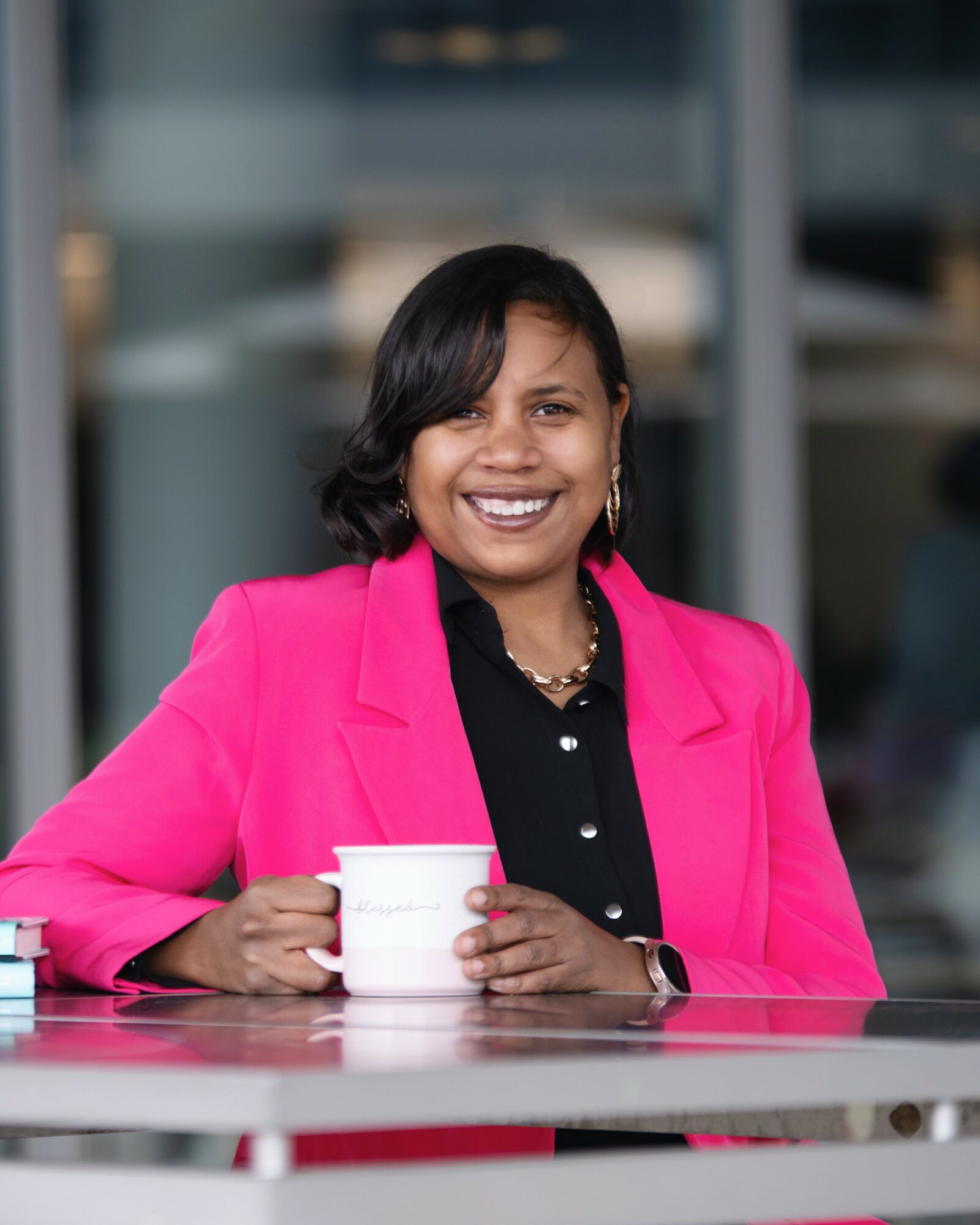 Woman smiling, wearing a pink blazer and black shirt, holding a white mug, sitting outdoors with a blurred background.