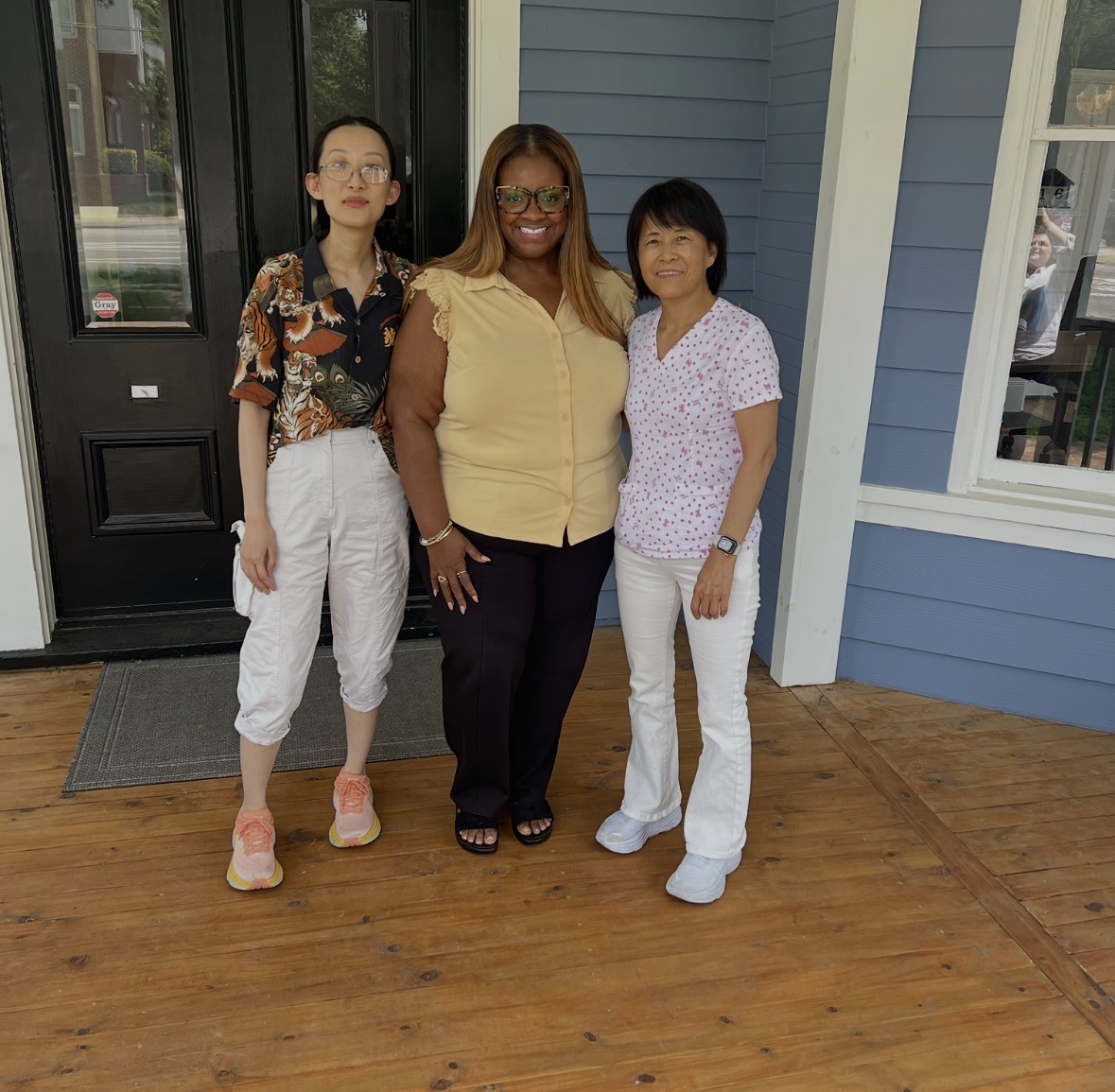 Three women standing on a wooden porch in front of a blue house, smiling at the camera.