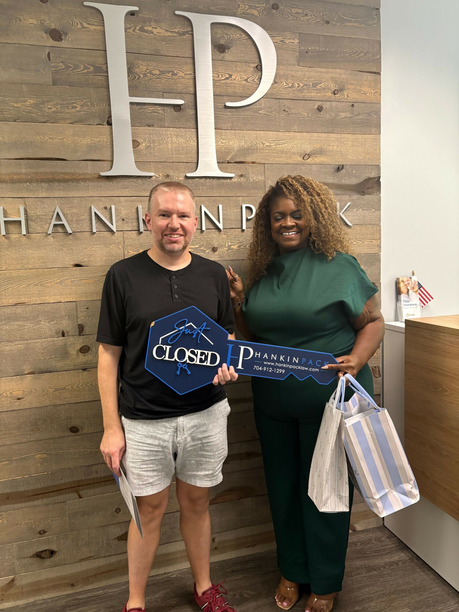 Two people standing indoors, one holding a diploma and a gift bag, with large decorative letters on the wall behind them.