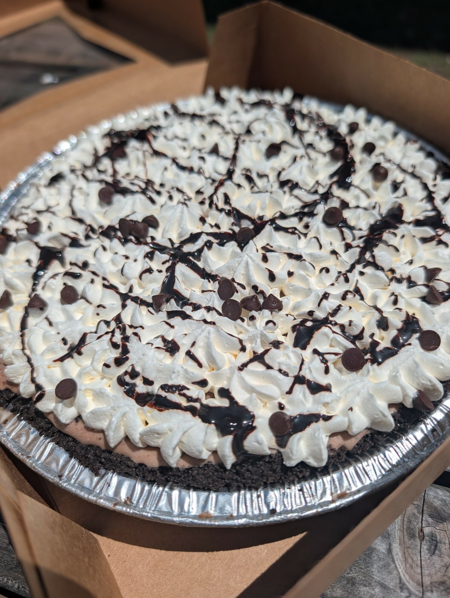 Chocolate cake topped with whipped cream and chocolate drizzle in a foil container, inside a cardboard box.