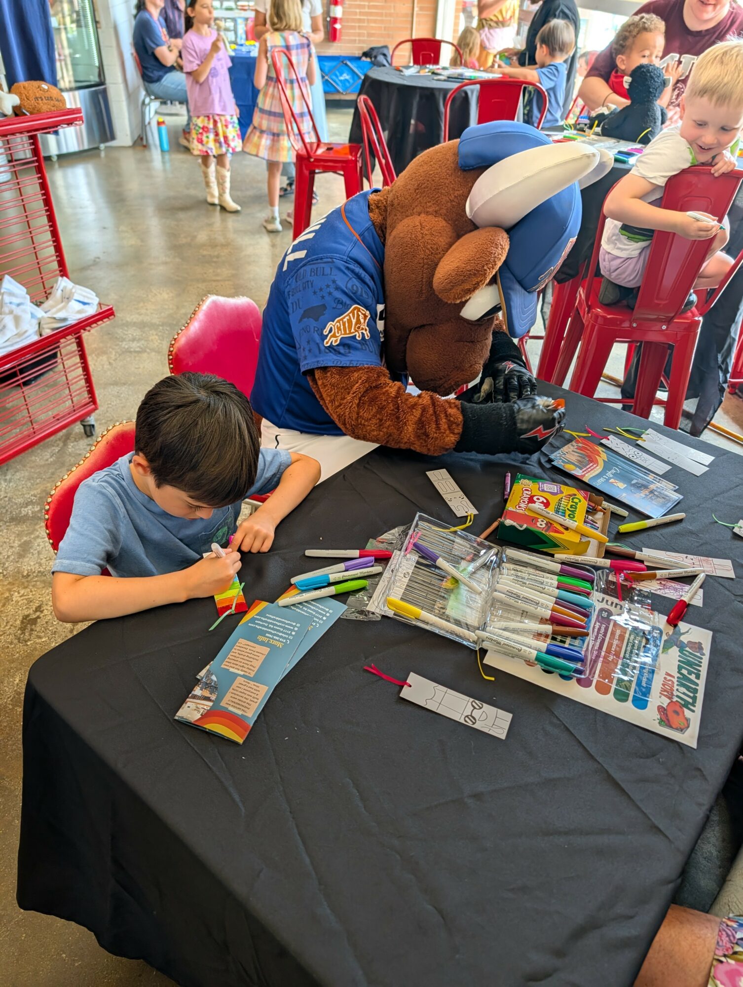 Child and person in mascot costume at table with art supplies, children in background, indoor setting.