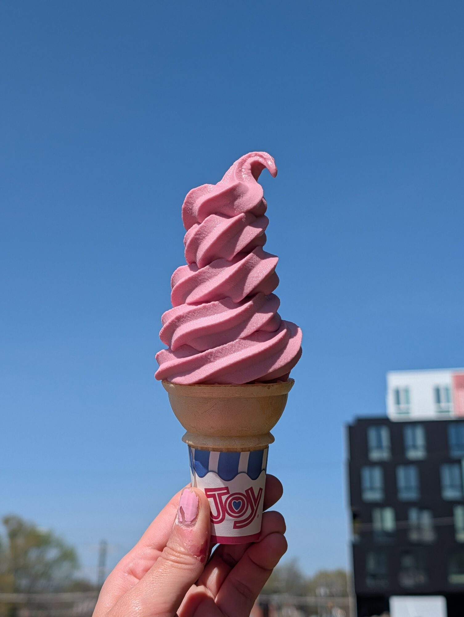 Hand holding a pink soft serve ice cream cone against a clear blue sky, with buildings in the background.