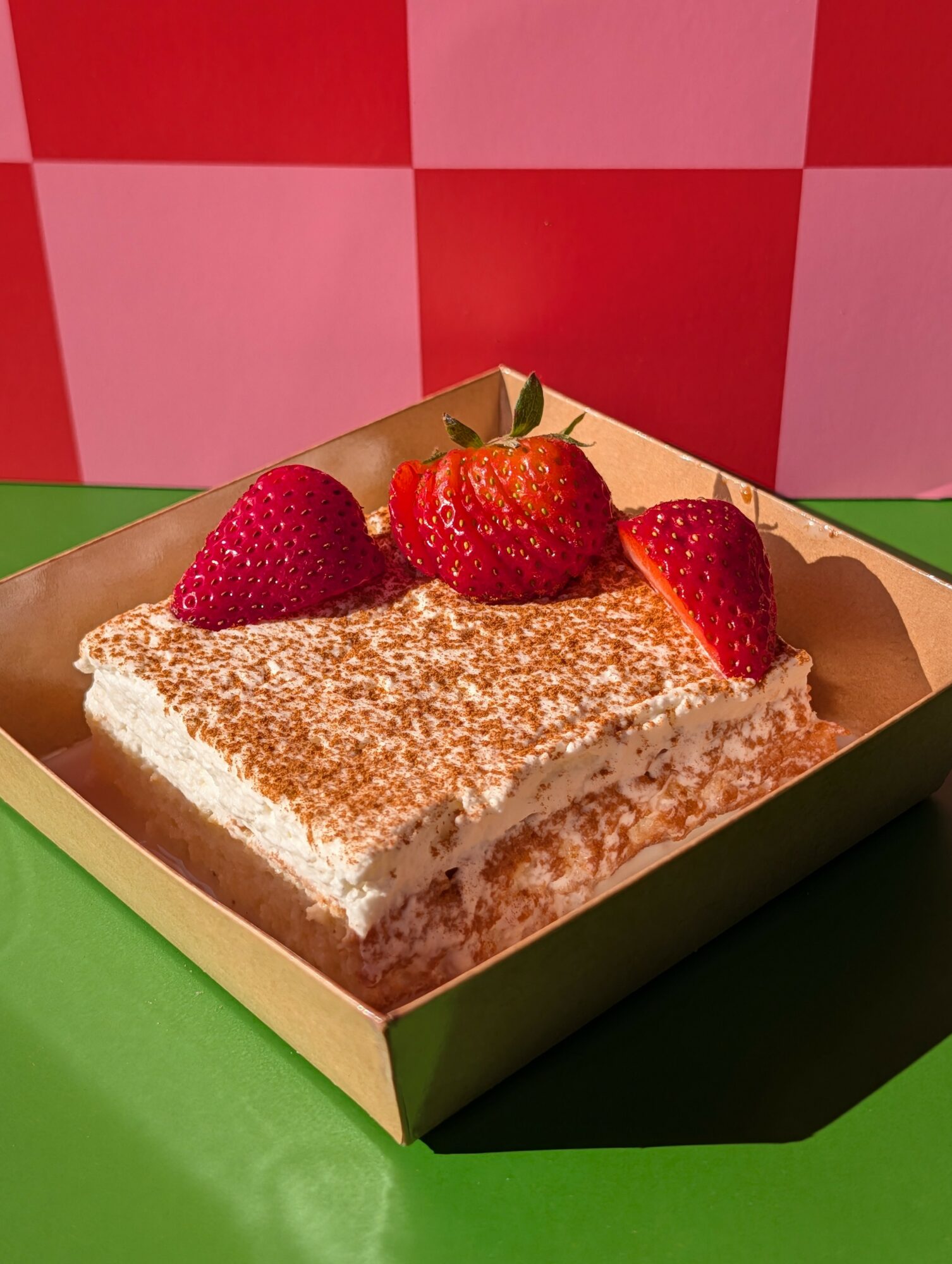 Square dessert topped with strawberries, dusted with powdered sugar, in a green tray on a green surface, with a red and pink checkered background.