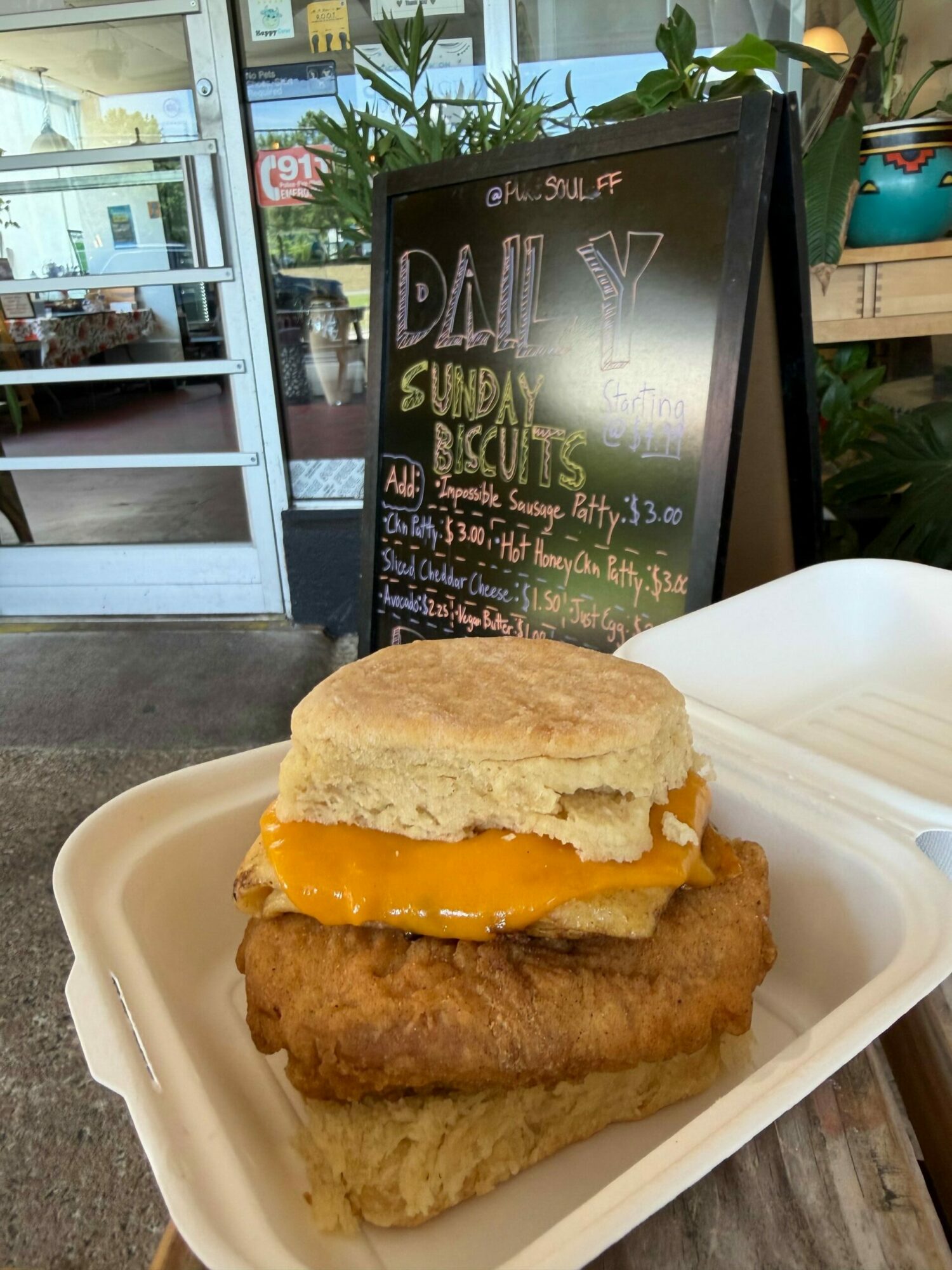 Fried fish sandwich with cheese in a bun, served with fried fish fillet on a white tray, in front of a chalkboard menu.