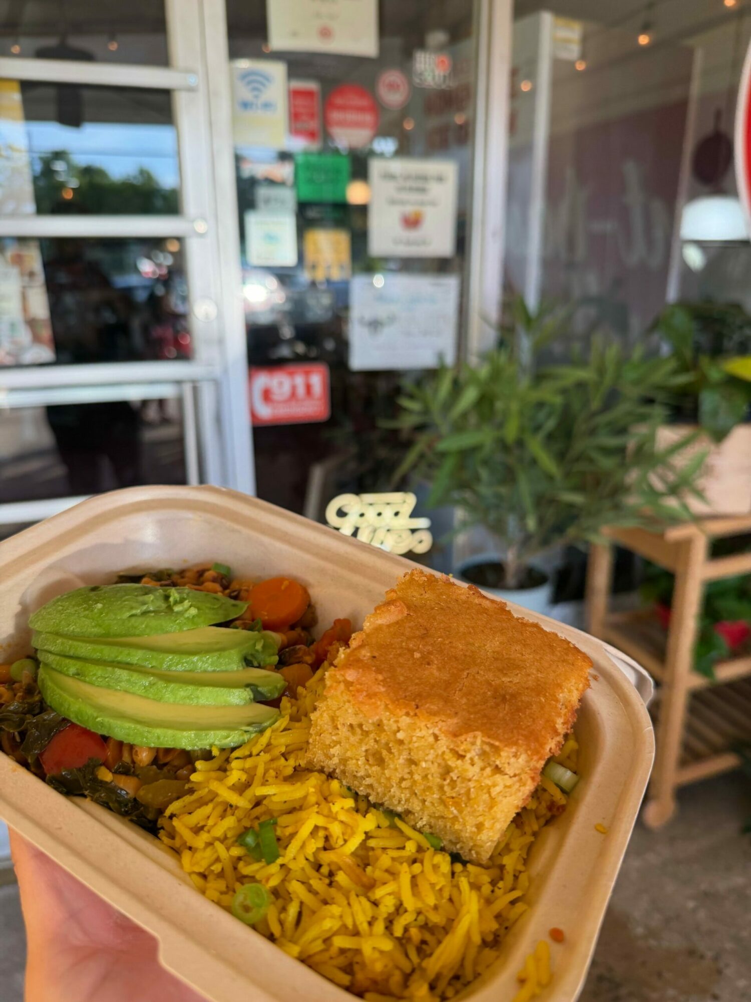 A takeout container with rice, fried food, and vegetables, held outdoors near a storefront with plants and signs.
