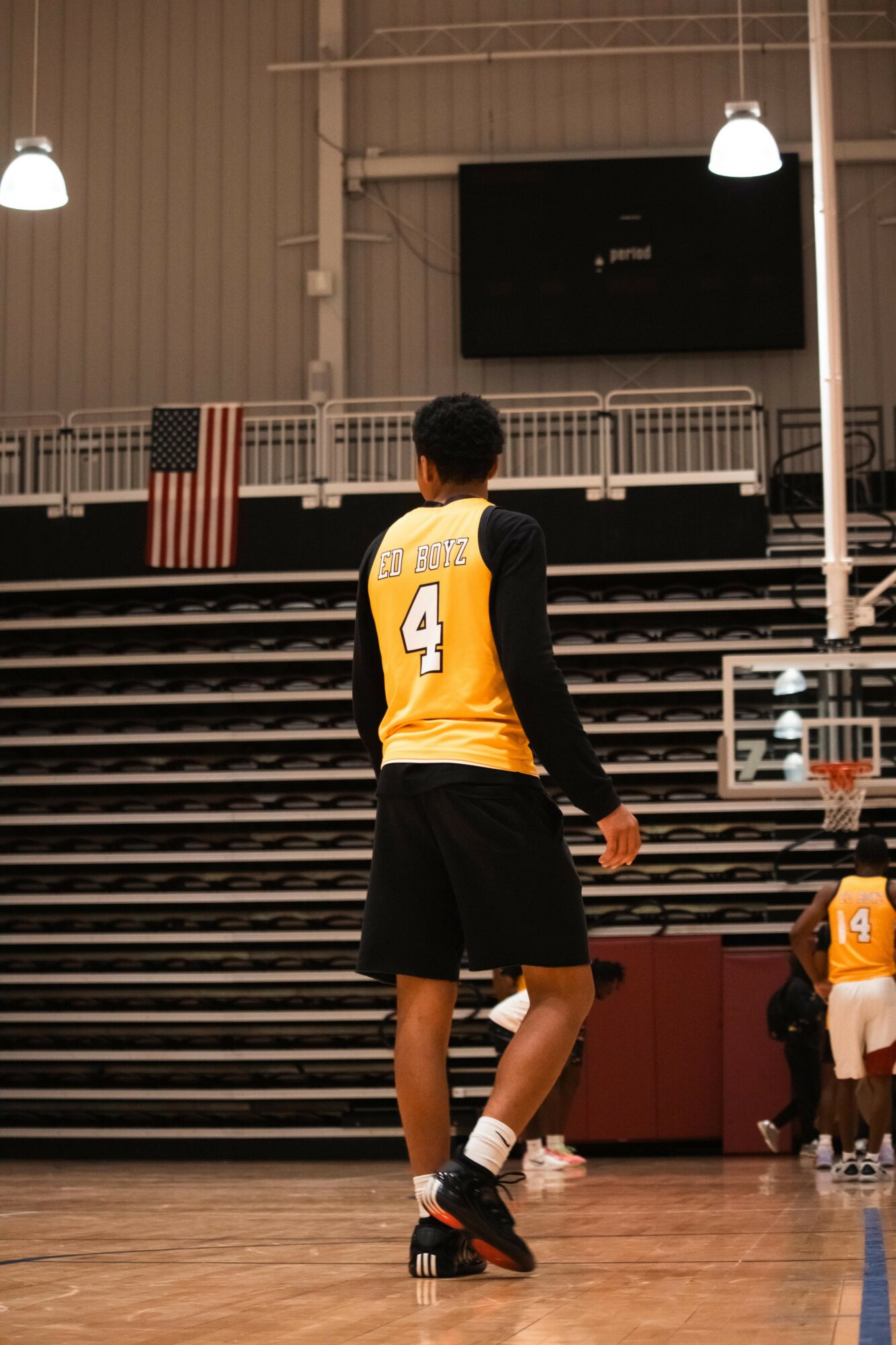 Basketball player wearing a yellow jersey with number 4, standing on the court facing away, with bleachers and a scoreboard in background.