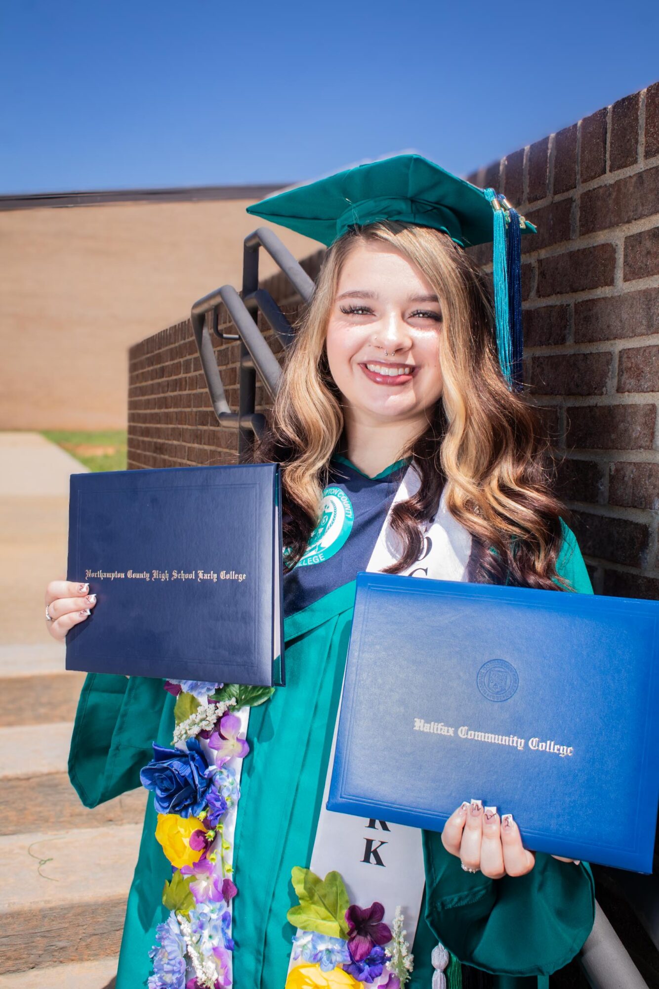 Young woman in graduation gown and cap holding diploma and certificate, smiling outdoors against brick wall.